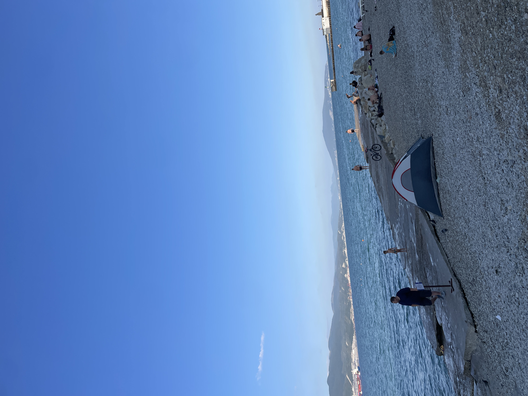 The image captures a scene at a rocky beach with the sea and a distant coastline under a clear blue sky. A man in a dark blue t-shirt and shorts stands on the paved area near the water, facing away from the camera. To his left, a small, partially set-up tent with red, white, and blue fabric is visible. A bicycle is leaning against the paved area near the tent.
Further along the paved walkway, several people are seen relaxing and sunbathing on the rocky beach. Some individuals are sitting or lying down, while others appear to be wading in the shallow water. The sea is calm with gentle ripples. In the background, a long pier or breakwater extends into the water, with structures at its end. The distant coastline features hills or mountains, and some buildings can be discerned along the shore.
The clear sky and bright lighting suggest it is daytime, likely late afternoon or early evening given the angle of the light. The presence of people in swimwear and engaging in beach activities indicates a warm, pleasant weather condition. The overall atmosphere appears relaxed and leisurely. No visible text is present in the image. The location is identified as Ivanovskaya, Russia.