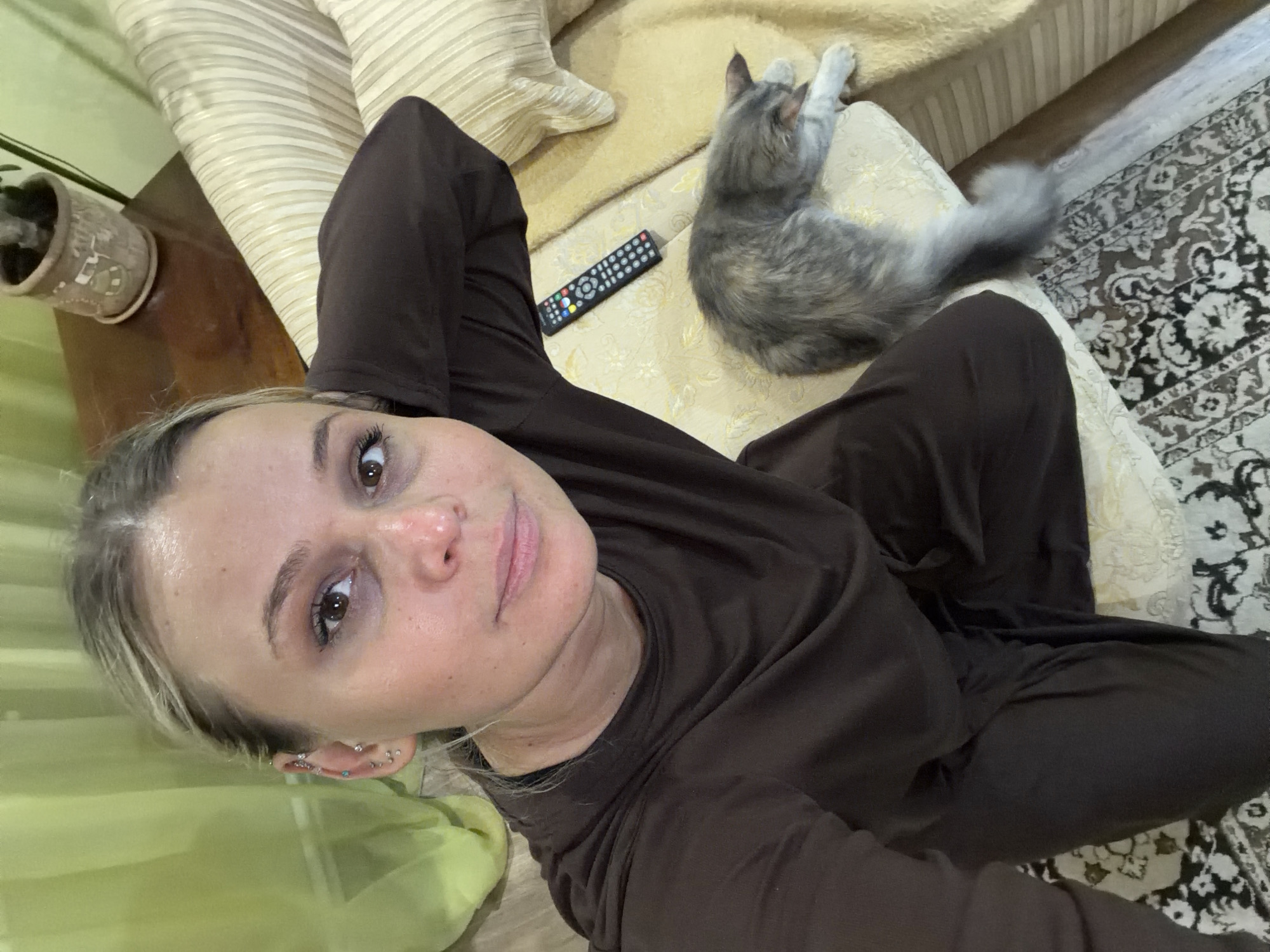 A woman wearing a brown long-sleeved shirt and pants is sitting on a beige couch with striped cushions. Next to her, on the couch, lies a gray cat. A black remote control is also on the couch, near the cat. Part of a rug with a patterned design is visible on the floor. A potted plant sits on a wooden surface near the woman. The picture was taken indoors, presumably in Sarapul, Russia.