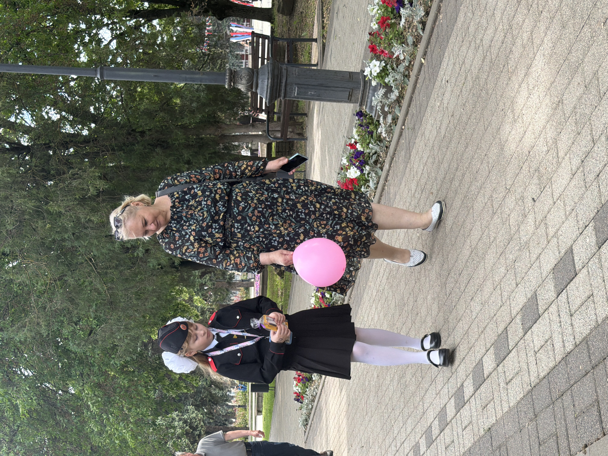 The image depicts a woman and a girl standing on a brick sidewalk in what appears to be a park. The woman is wearing a floral-patterned dress, while the girl is wearing a dark uniform with white tights and black shoes. The girl is holding a clear container of a brownish liquid. The woman is holding a pink balloon. A black lamppost and flower beds are visible in the background. Based on the available information, it is impossible to confirm that the location is Leningradskaya, Russia. The image suggests it is an outdoor public space.