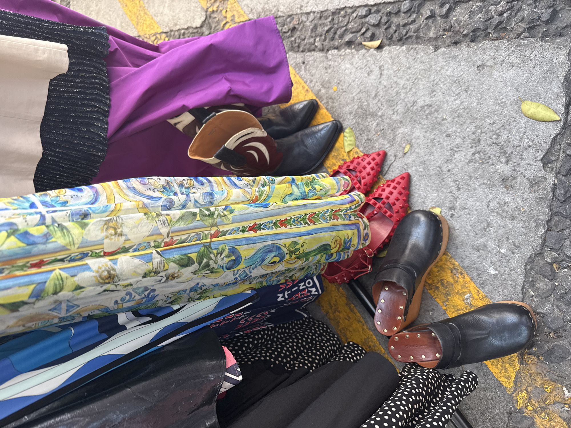 The image shows a collection of items on a concrete surface marked with yellow stripes, suggesting a street in Mexico City, Mexico. The items include a pile of folded fabrics in various patterns, colors, and materials, such as a brightly colored floral print, a black and white polka dot pattern, a solid purple fabric, and other striped or patterned fabrics. There are also several pairs of shoes: a pair of black leather cowboy boots, a pair of red studded shoes, and a pair of black leather clogs with wooden soles. The items are scattered and piled on top of each other, indicating a temporary or informal arrangement. The surrounding environment is plain concrete, with a few scattered leaves, hinting at an outdoor setting.