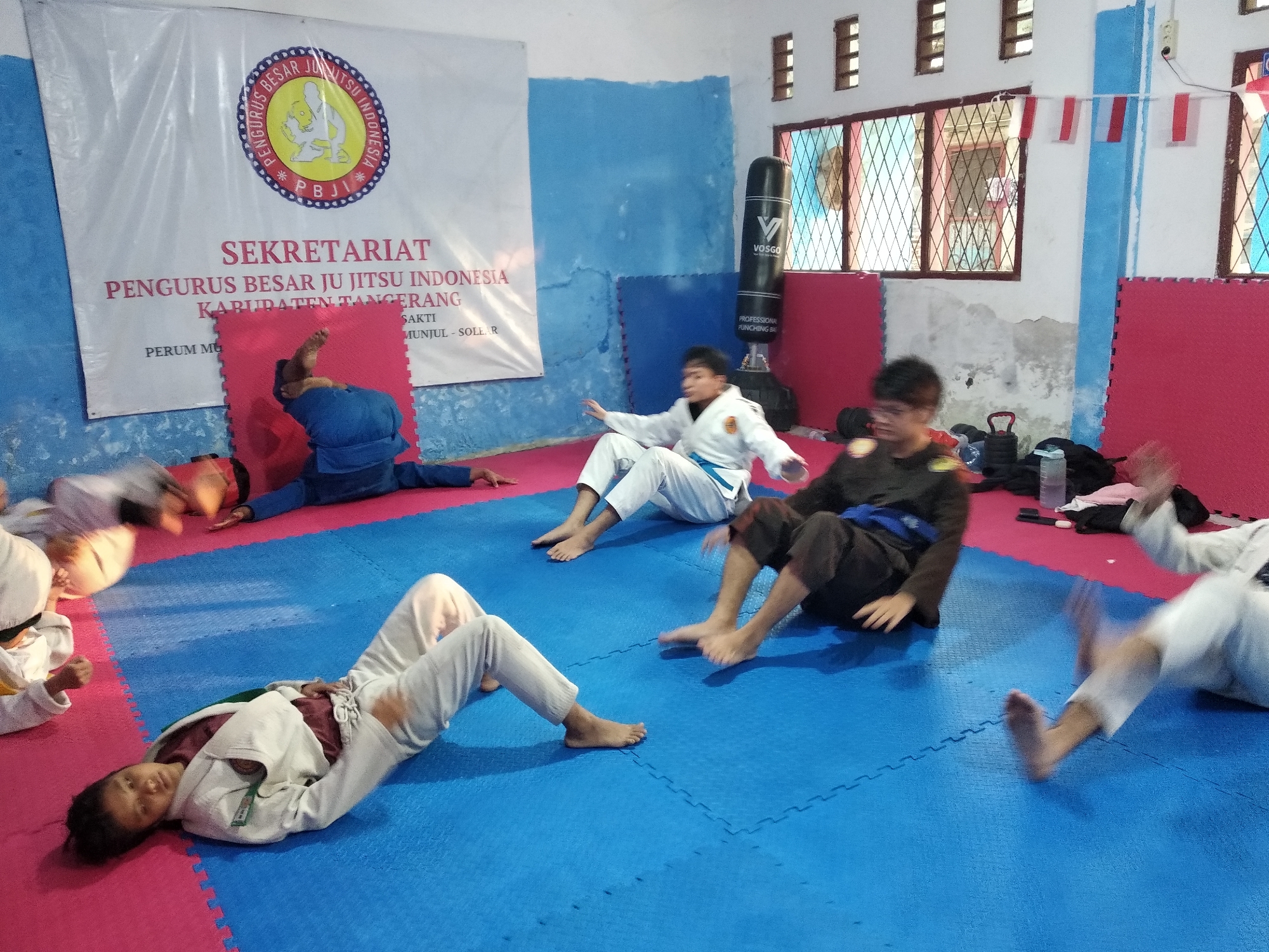 The image depicts a martial arts training session, likely in a gymnasium or dojo, located in Munjul, Indonesia. The floor is covered with blue and pink interlocking mats. In the background, a large white banner with red text and a circular logo hangs on a blue wall. The banner appears to be for an organization named "PENGURUS BESAR JU JITSU INDONESIA KABUPATEN TANGERANG" with the location "MUNJUL - SOLE.R" inscribed on it.
Several individuals are engaged in various activities related to jiu-jitsu training. In the foreground, one person in a white gi is lying on their back on the blue mat, looking towards the camera. To the left, another individual in a white gi is also on the mat, with their upper body partially visible. Further back, a person in a blue gi is positioned on the floor, with their legs raised.
In the mid-ground, two individuals are seated on the mat. One, wearing a white gi, is looking to the right with their hands outstretched. Another, in a dark gi with a blue belt, is also seated, with their legs extended. To the right, another person in a white gi is in motion, possibly demonstrating a technique, with their legs blurred due to movement.
Additional objects in the scene include a black punching bag with the logo "VOSGO" visible, situated against the blue wall. Several Indonesian flags are displayed on the wall near a window. Other items such as weights, water bottles, and bags are scattered around the periphery of the training area. The overall impression is one of a dynamic and active martial arts practice environment.