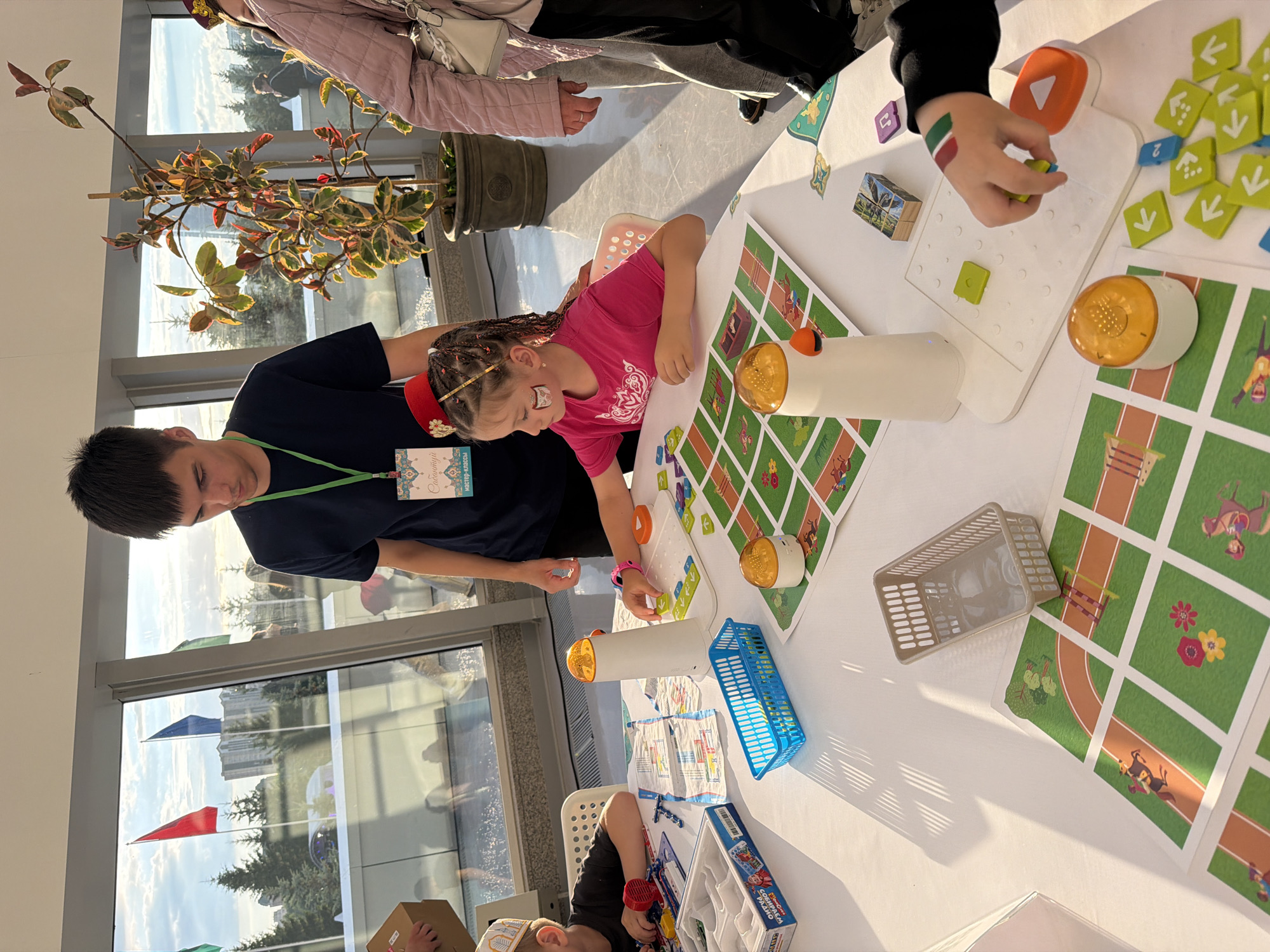 A young girl in a pink shirt and a red hat with gold trim is playing a game at a white table, her face adorned with face paint. She is positioned on the right side of the frame, her left hand engaged with a white electronic game board featuring green and blue directional arrows. To her left, a white tower-like device with an orange top sits on the table, adjacent to a game board with a racetrack motif. A young man wearing a navy blue t-shirt and a lanyard with a tag stands behind the girl, looking down at the game. He is positioned on the left side of the frame. The table is cluttered with various game pieces, including colorful tiles, a blue plastic basket, a grey plastic basket, and a boxed toy. In the background, large windows offer a view of trees, buildings, and flags, suggesting an outdoor setting or a venue with a scenic view. The lighting indicates it is daytime, possibly late afternoon, with sunlight casting shadows on the table.