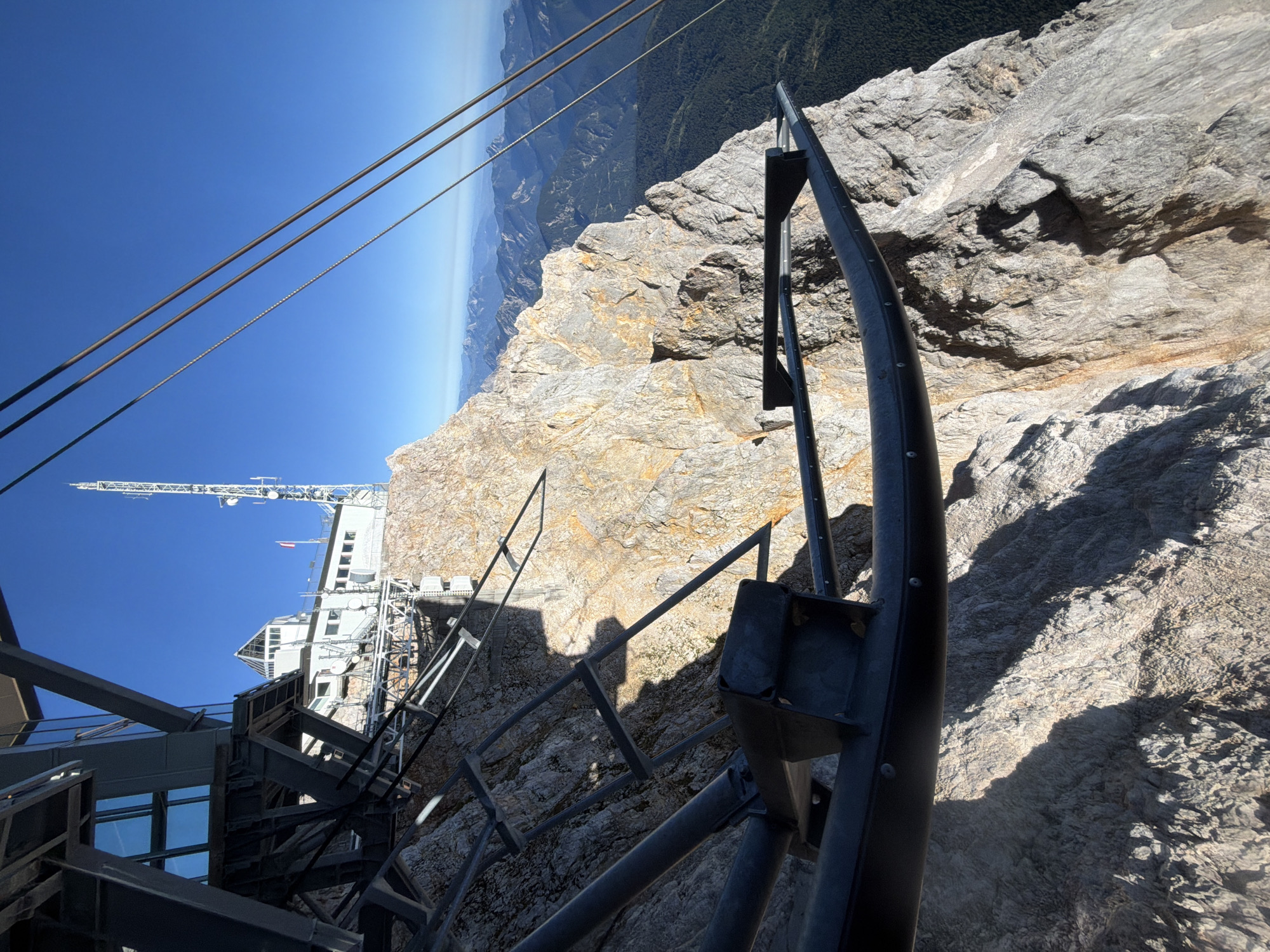 A high-angle, close-up shot captures a section of a cable car structure, including metal support beams and cables, positioned against a dramatic backdrop of the Zugspitze mountain in Grainau, Germany. The mountain's rocky, rugged terrain dominates the lower half of the frame, with a sheer drop visible. In the mid-ground, a white building with antennas and a telecommunications tower is perched on the mountain peak. The sky is a clear, vibrant blue. The composition emphasizes the scale and steepness of the mountain and the engineering marvel of the cable car system.