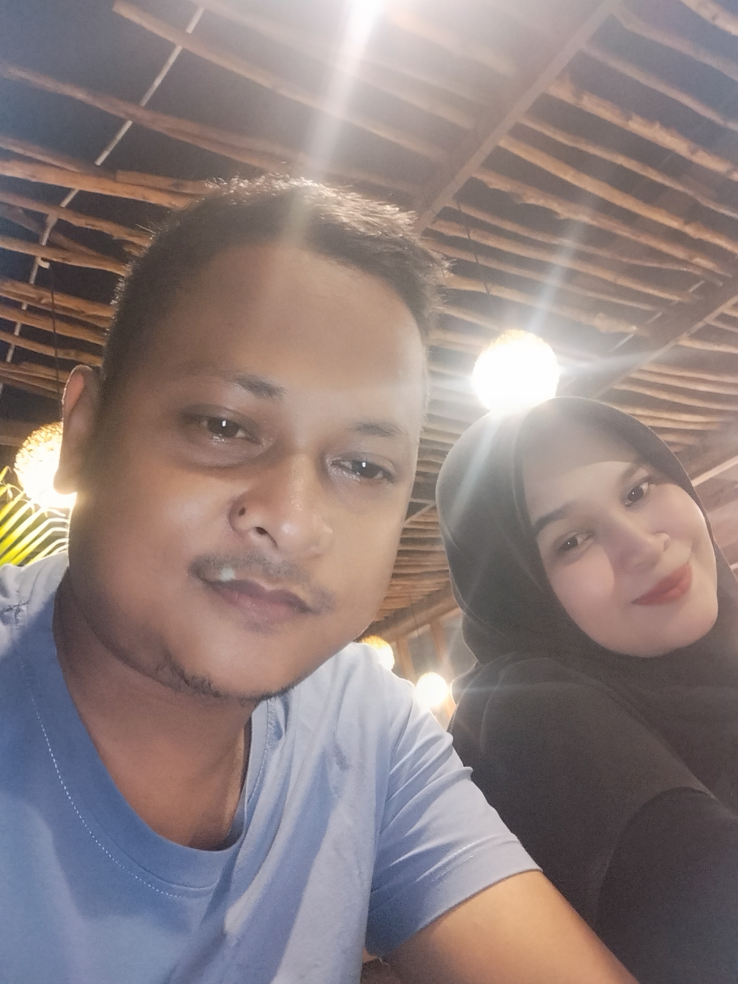 A selfie captures a man and a woman in what appears to be an indoor or covered outdoor setting during the evening or nighttime. The man, positioned in the foreground and occupying the left side of the frame, has short dark hair, a light mustache, and a small goatee. He is wearing a light blue t-shirt and looks directly at the camera with a neutral expression. To his right, partially behind him, is a woman wearing a black hijab and black clothing. She has subtle makeup, including red lipstick, and offers a gentle smile towards the camera.
The background features a distinctive ceiling structure made of light-colored wooden beams, creating a patterned effect. Several bright light sources, possibly decorative lamps, are visible, casting warm glows and noticeable lens flares across the upper portion of the image. These lights contribute to the ambient, cozy atmosphere of the location, which could be a cafe, restaurant, or similar establishment in Keudekruenggeukueh, Indonesia. The overall mood conveyed by the subjects is relaxed and content. There is no visible text in the image.
