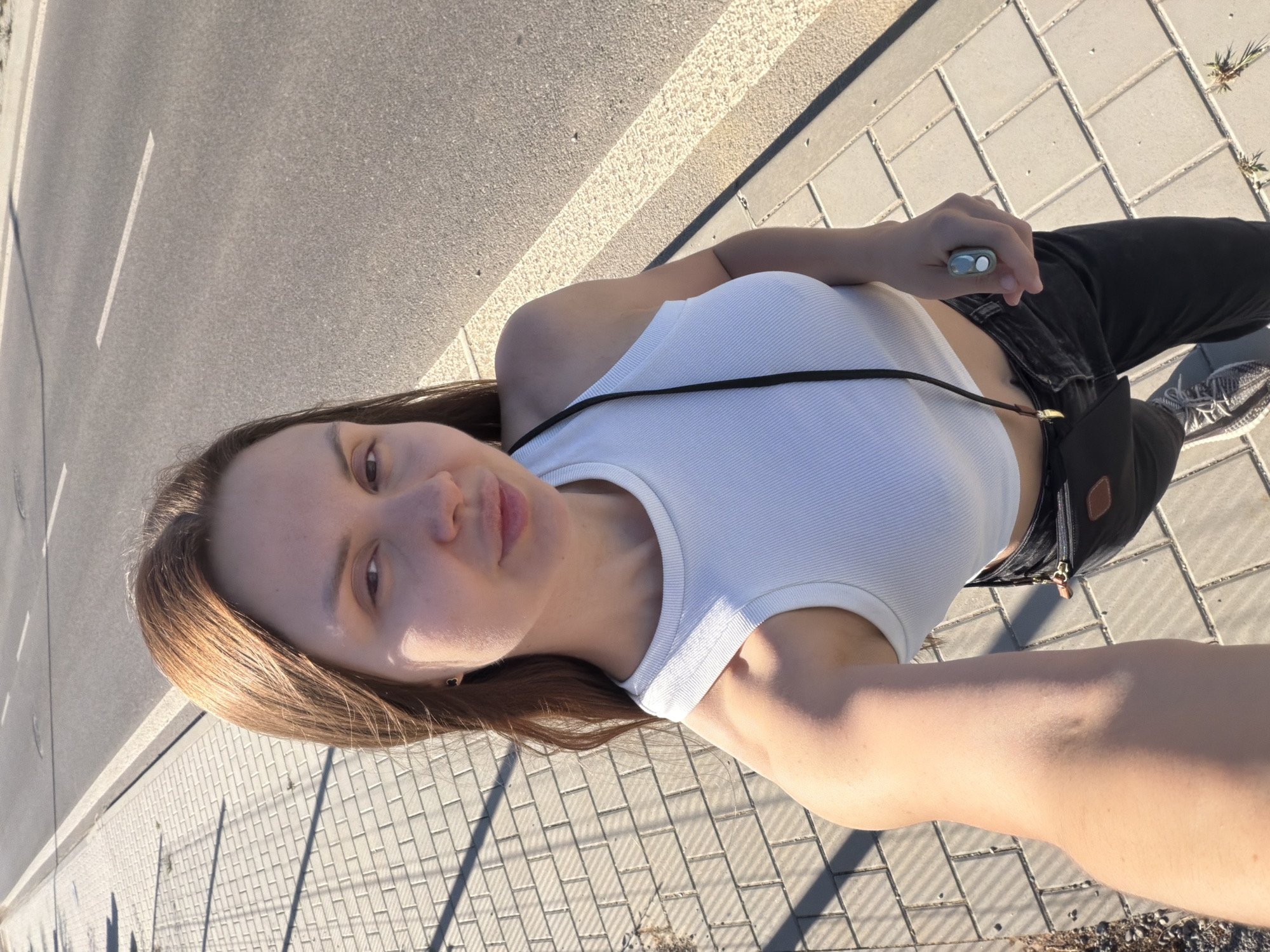 A woman with long brown hair is taking a selfie outdoors. She is wearing a white ribbed crop top, black pants, and gray sneakers. She has a small black purse slung over her shoulder and is wearing a ring on her right hand. The woman is standing on a paved sidewalk next to a road with a yellow painted line. The sun is casting shadows on the sidewalk. The image appears to have been taken in the city of Nitra, Slovakia.