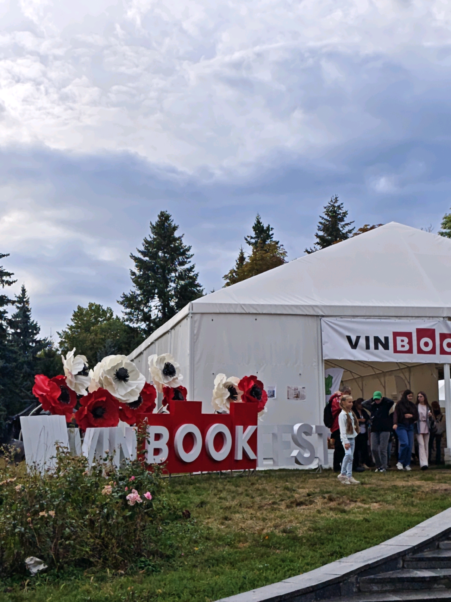 The image depicts a scene outside a large white tent with a banner that reads "VINBOOK" on the front. In front of the tent, there is a display of large red and white paper flowers. To the left of the flowers, there are large white letters spelling out "VIN BOOK FEST". Several people are visible standing and walking in front of the tent entrance, some of them appearing to be attendees. The environment appears to be an outdoor setting with trees in the background and grass in the foreground. The scene is likely from the Vinnytsia Book Festival, held in Vinnytsia, Ukraine.