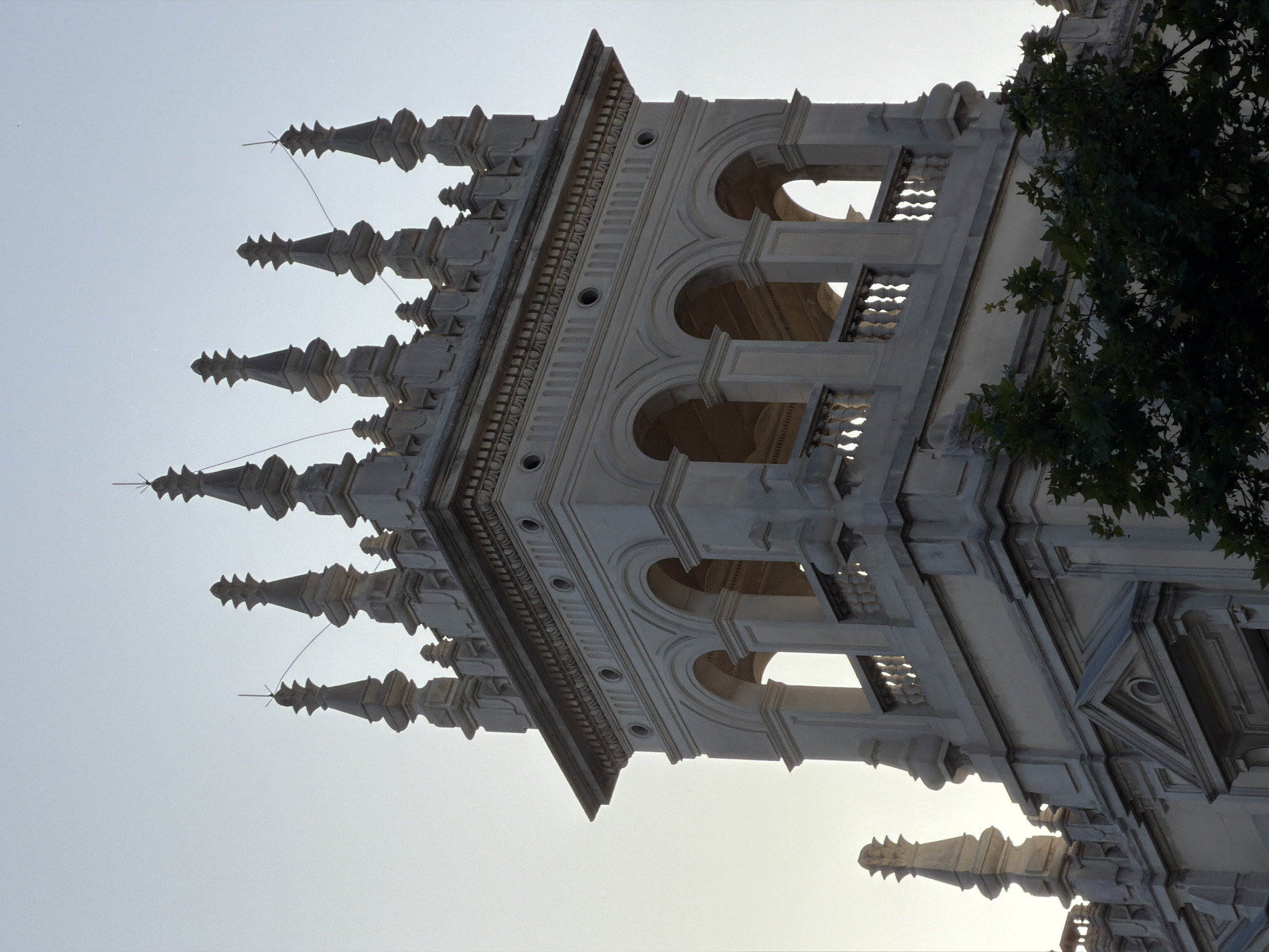 The image captures a detailed, upward-looking view of an ornate, light-colored stone building, likely a monumental structure or cathedral, set against a bright, clear sky. The perspective emphasizes the towering height and intricate craftsmanship of the architecture.
The structure features numerous pointed spires or finials, each topped with a small, delicate spike, extending along the left side of the frame and tapering upwards. These spires are connected by thin, almost invisible wires. Below these spires, the building showcases multiple levels of arched openings, forming what appear to be balconies or decorative windows. These arches are supported by elegant balustrades with finely carved details. The stone surfaces between these architectural elements are adorned with rich moldings, including a series of small, circular holes and vertical fluting.
A tree with dark green foliage partially obscures the upper right section of the building, providing a natural contrast to the pale stone. The sky is a uniform, bright grey-blue, suggesting the time of day is either early morning or late afternoon, as sunlight gently illuminates the building from the side, creating soft shadows that enhance its three-dimensional quality. No people are visible, and there is no discernible activity taking place. The image focuses solely on the static beauty and intricate details of the architecture. No text is visible. Given the location context of Barcelona, Spain, the architectural style is indicative of historic Gothic or Neo-Gothic structures in the city, such as the Barcelona Cathedral.