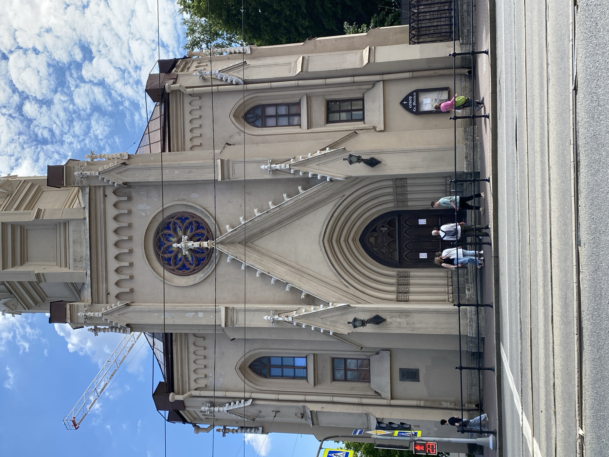 The image displays a daytime street scene in Saint Petersburg, Russia, centered on the facade of a grand, light-colored building, identified by a sign as the "СОБОР Св. МИХАИЛА" (Cathedral of St. Michael). The cathedral exhibits Gothic Revival architecture with pointed arched windows, intricate stone tracery, decorative finials, and a prominent rose window featuring blue and gold stained glass.
The building is situated alongside a city street with tram tracks and a wide sidewalk, separated by black metal railings. Several individuals are present on the sidewalk: two people are near the main arched entrance, another person in a light blue top stands nearby, and a person in a pink top and green backpack is further down the sidewalk near a dark sign. In the foreground, another person in a dark top is visible near a pedestrian crossing, where a traffic light shows a red signal with the number "23" counting down.
The sky is bright blue with scattered white clouds, indicating clear and sunny weather. A large construction crane is visible on the left side of the frame, extending upwards beside the cathedral. The scene depicts a typical urban environment with historical architecture and daily pedestrian activity.