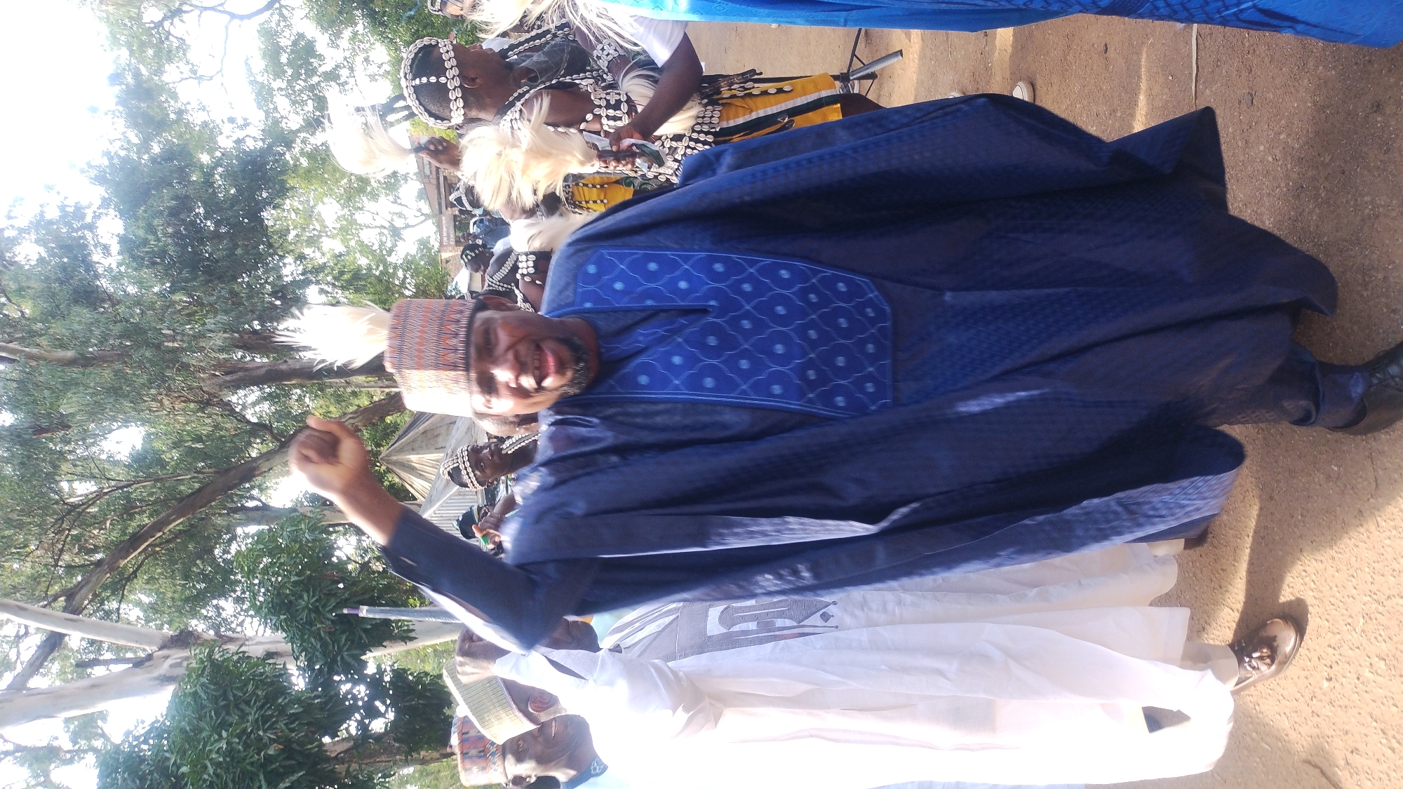 The image depicts a man in a vibrant blue traditional outfit, including a flowing gown and a patterned cap, raising his fist in the air. He appears to be smiling and engaged in a lively event, possibly a celebration or a cultural gathering. He is surrounded by other individuals in similar traditional attire, some of whom are adorned with decorative necklaces and headdresses. The background suggests an outdoor setting with trees and what appears to be a dirt or sandy ground. Based on external knowledge, the image is believed to have been taken in Jos, Nigeria.