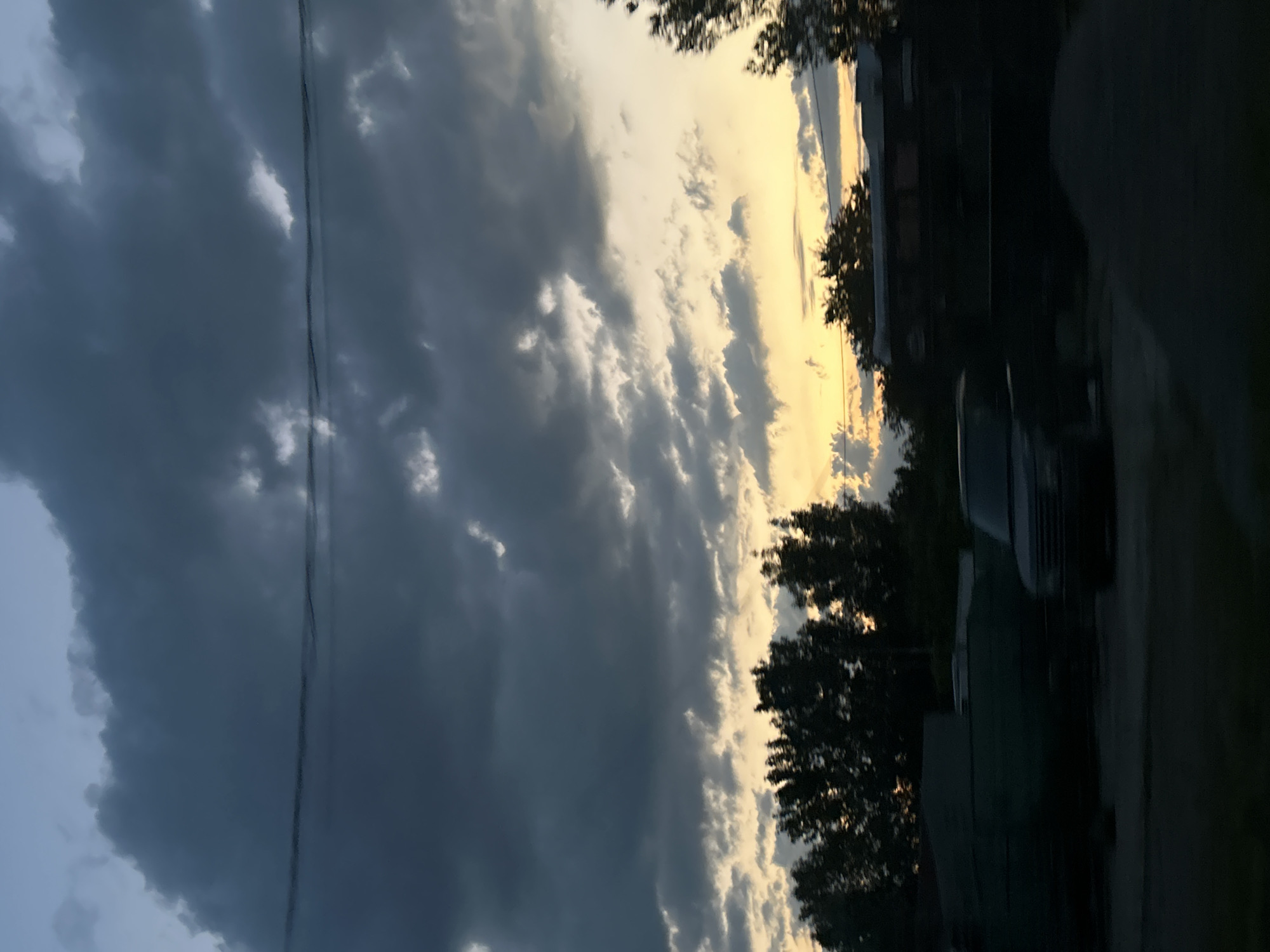The image shows a blurred view of a residential area at dusk. Dark, ominous clouds fill the sky, with streaks of yellow and orange light peeking through the gaps, suggesting the sun is setting or has recently set. Silhouetted trees and structures are visible in the mid-ground, including what appears to be a dark-colored SUV or similar vehicle in motion on the right side of the frame. The foreground is also dark, with indistinct shapes that could be foliage or debris. The overall impression is one of a moody, possibly stormy evening in a semi-urban or rural setting.
