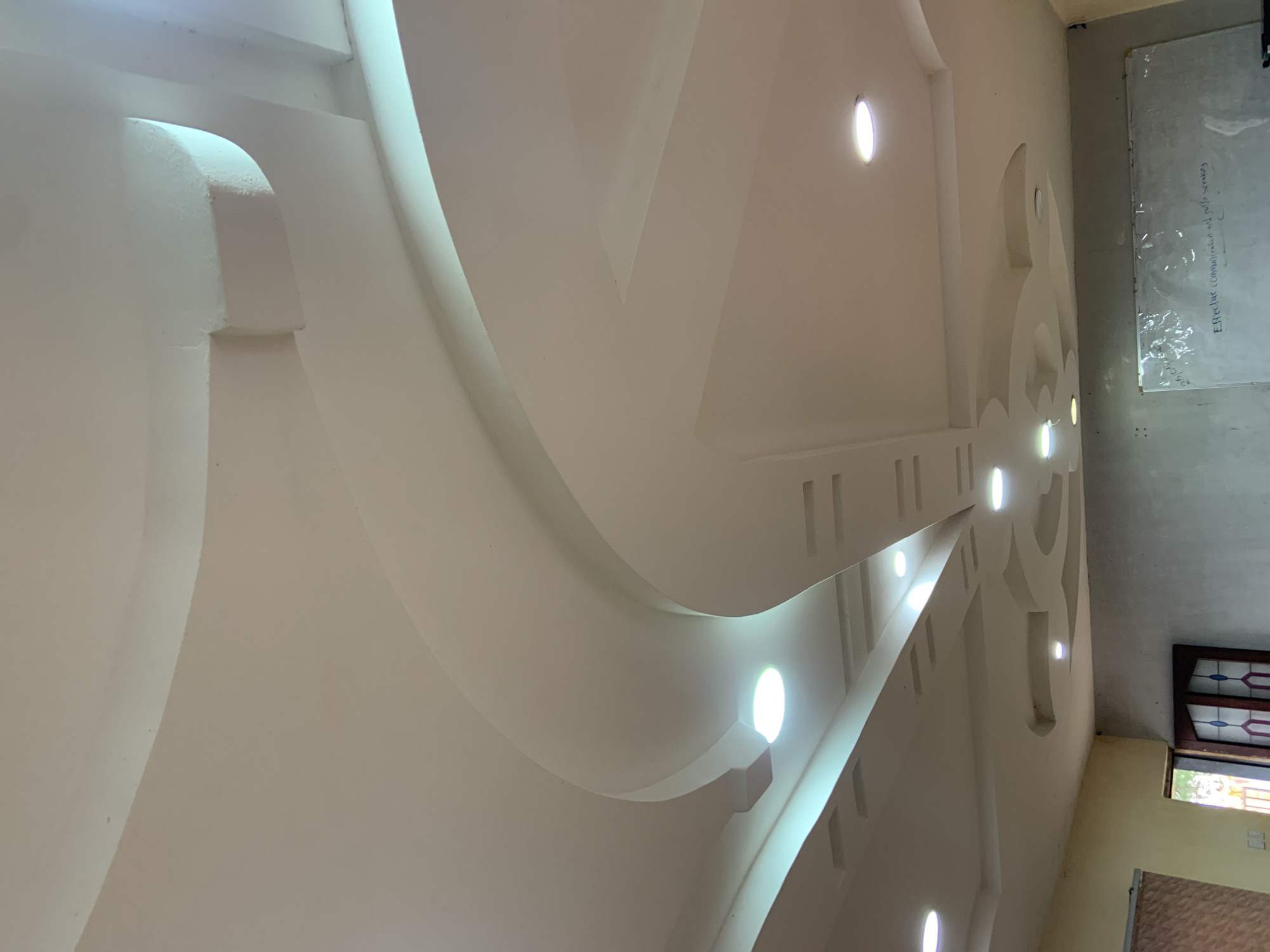 The image shows a detailed view of a modern, multi-layered ceiling with integrated lighting. The ceiling features curved designs and recessed sections, illuminated by several circular spotlights. The lighting casts a soft glow across the textured surfaces. In the lower part of the image, a whiteboard is visible, with some text written on it. To the left, a partially open doorway reveals a glimpse of the outdoors, and a brown-framed window with textured blinds is also visible. Adjacent to the doorway, a wooden door with decorative glass panels is present. The walls appear to be painted in neutral tones. The overall impression is of an indoor space, possibly an office or meeting room, characterized by contemporary interior design. There are no people or animals visible in the image. The location is identified as Gura Top, Nigeria.