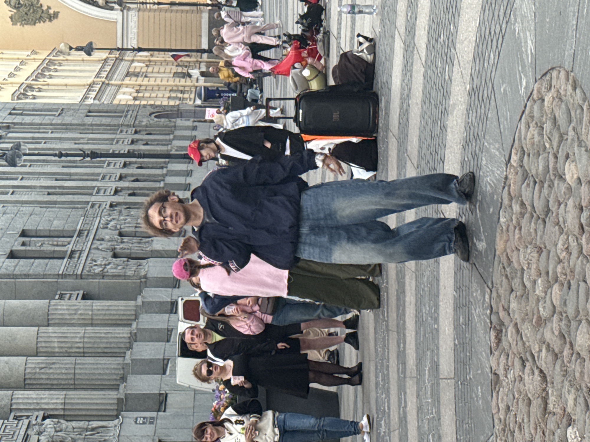 A crowd of people are gathered on a paved plaza with cobblestone sections in front of a neoclassical building in Saint Petersburg, Russia. The building features large stone columns and decorative relief sculptures. In the foreground, a man with curly hair and glasses is wearing a dark blue jacket and loose-fitting blue jeans. He is gesturing with his right hand. Behind him, a variety of individuals are visible, including a woman in a black dress, a woman in a pink hat and pink sweatshirt, and others in casual attire. Several pieces of luggage, including a large black JBL speaker on wheels, are present on the ground. The overall scene suggests a public gathering or event.