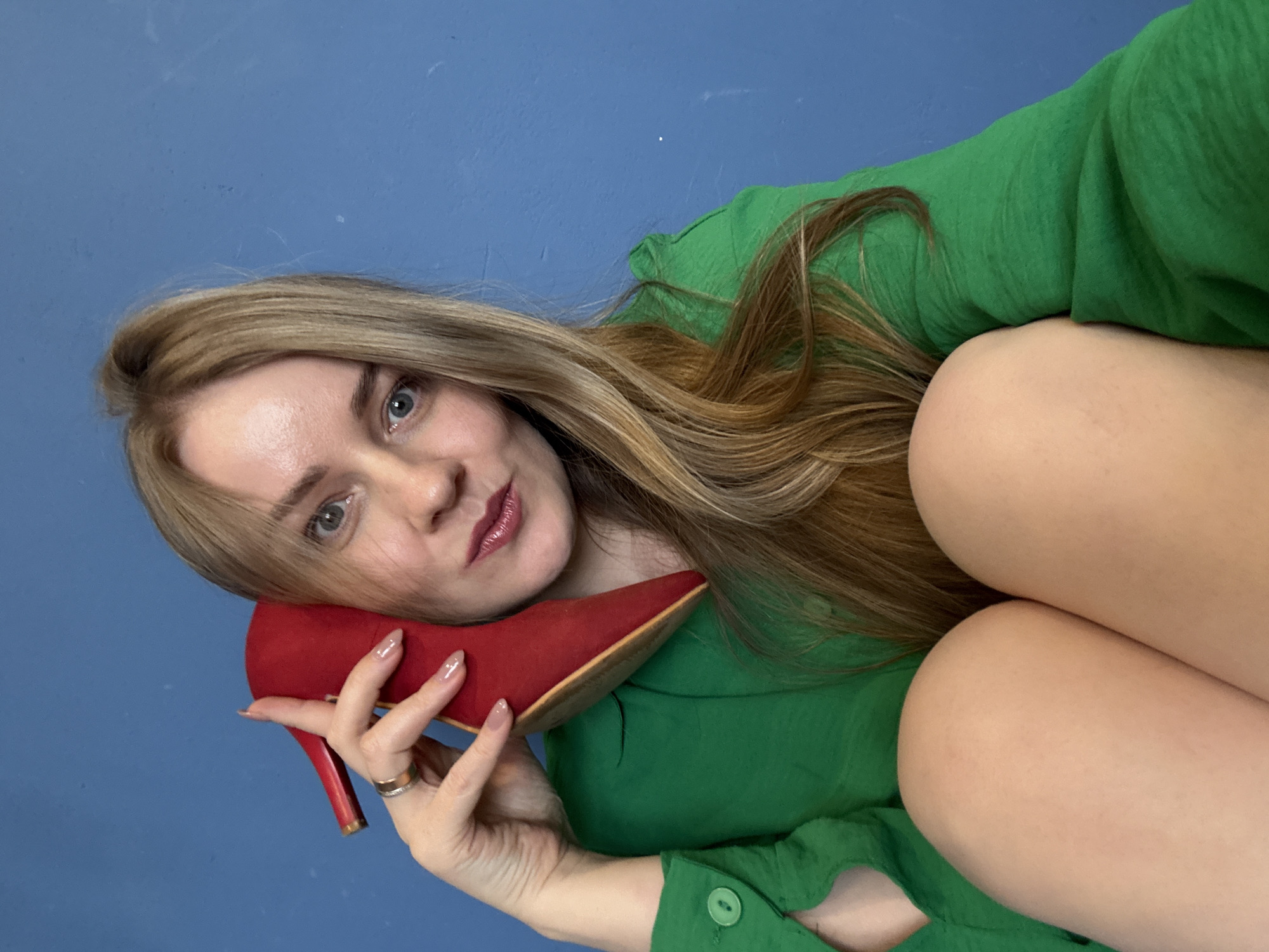 A fair-skinned woman lies on a blue surface, possibly a floor or wall, with her head tilted to the left. She has light brown hair with highlights, fair skin, and light eyes. She wears a green button-down shirt. She holds a red high-heeled shoe against her face. She wears a ring on her left ring finger. Her legs are visible, bent at the knees and drawn towards her torso. The location is indeterminate. There is no information available about the city of Chaikovsky, Russia.