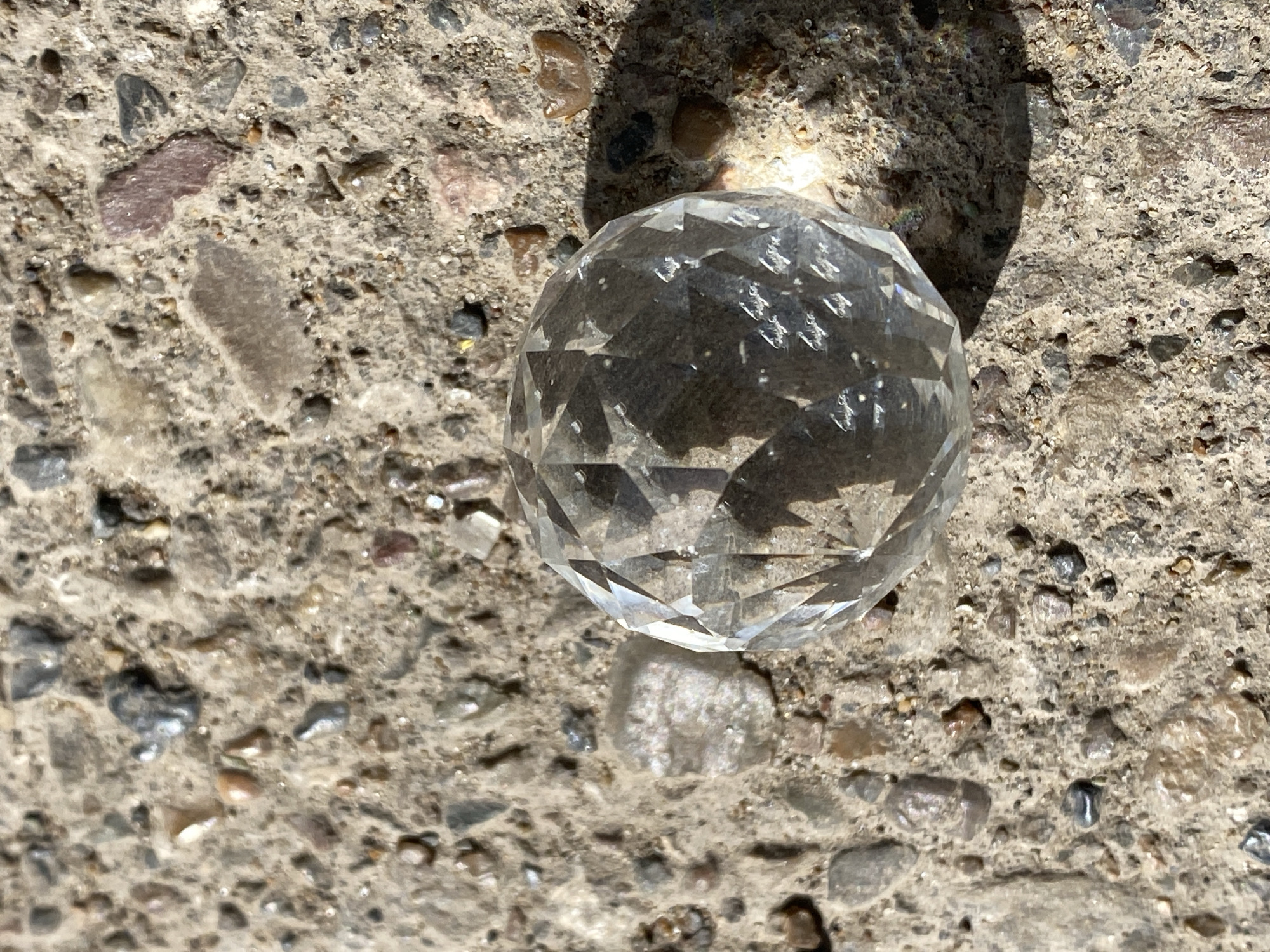 A multifaceted clear glass sphere rests on a rough, concrete surface with embedded pebbles and stones. The sphere is positioned slightly to the right and center of the frame. A shadow of the sphere and possibly a portion of the surface it rests on is visible behind it. The concrete surface is composed of various sizes and colors of pebbles and stones, including gray, tan, and reddish hues. The scene is lit by direct sunlight, casting shadows and highlighting the textures of the concrete and the facets of the glass sphere. The location is not identifiable as Chaikovsky, Russia, from the visual information provided.