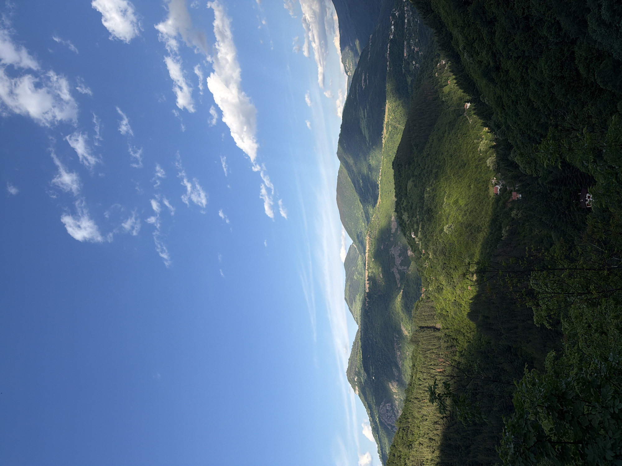 The image shows a landscape in Zasele, Bulgaria. There are green mountains with some houses nestled within the trees. The sky is blue with scattered white clouds. The sun is shining, casting shadows on the mountainside.