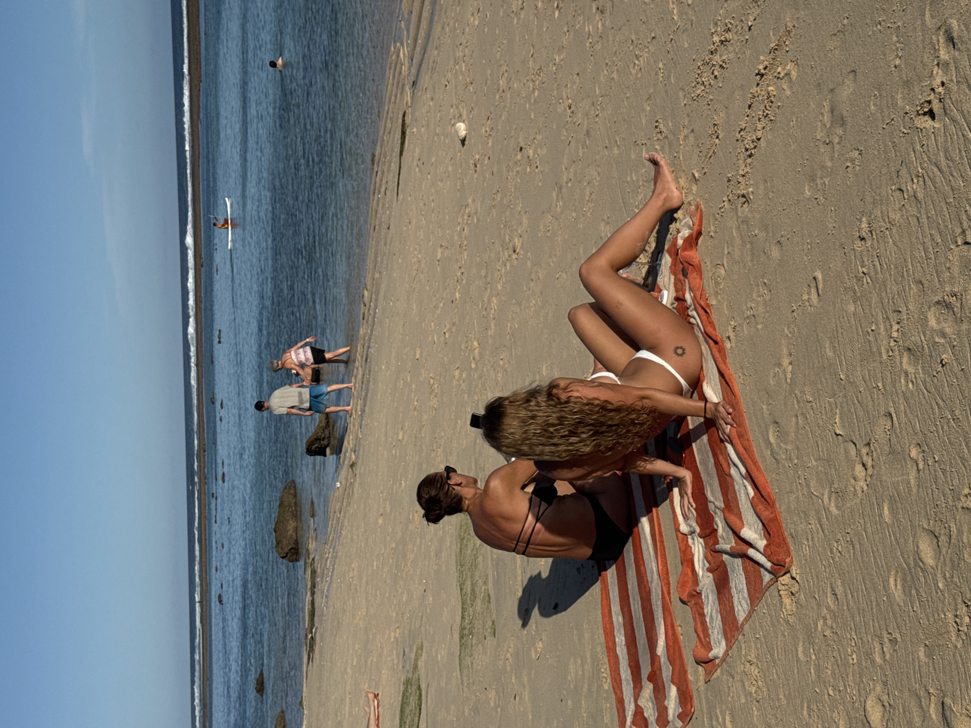 A full shot depicts a beach scene at Kuta Selatan, Indonesia. In the foreground, two women lie on a striped towel placed on the sand. One woman wears a white bikini and has a tattoo on her thigh. The other woman wears a black bikini and sunglasses.
In the middle ground, a man and a woman stand at the edge of the water, with another man standing further out, all near some rocks. In the background, a person is on a surfboard in the water, with another person swimming further out. The sky is clear and blue.