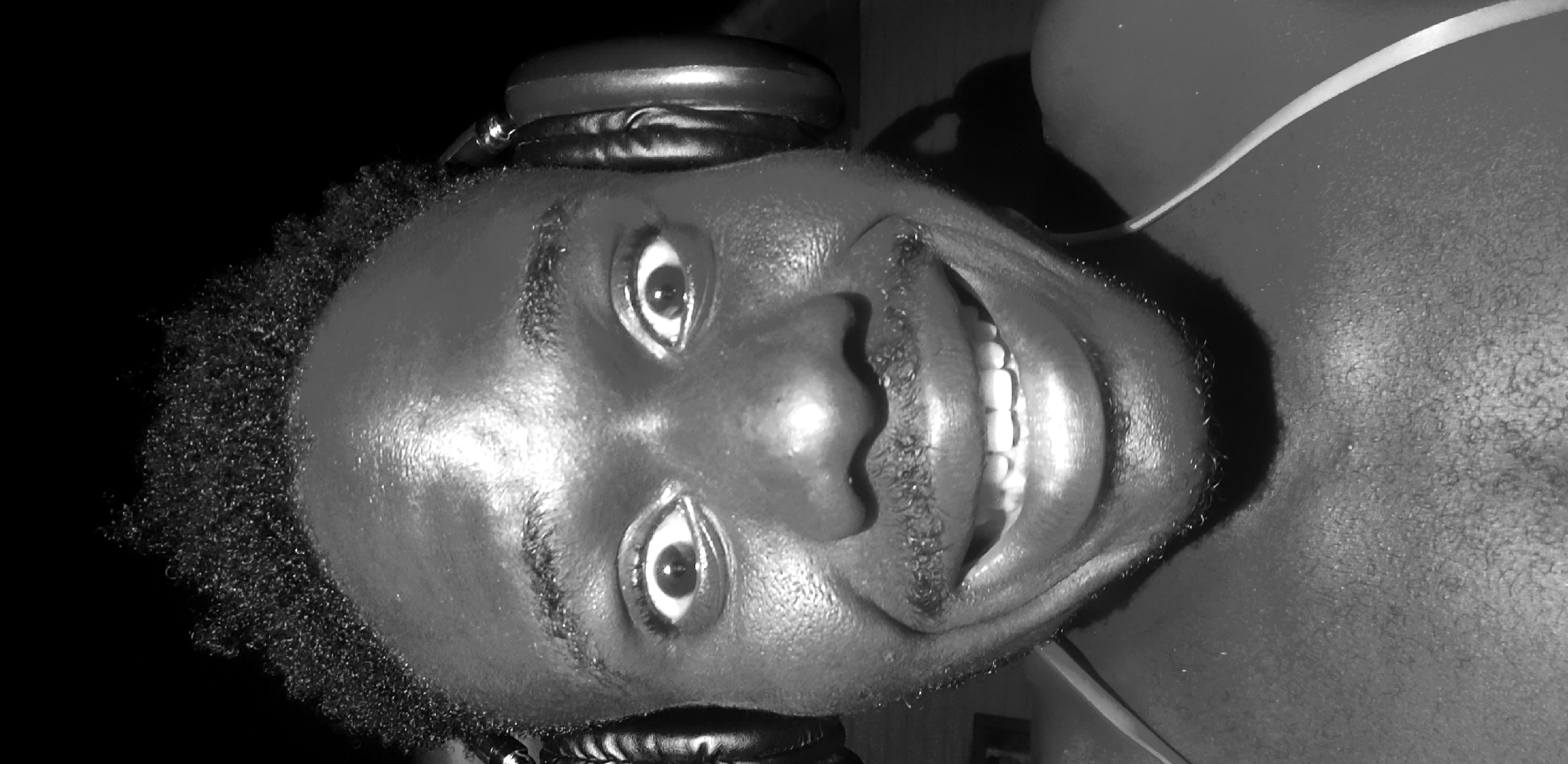 A close-up, black and white, selfie-style photograph shows a Black man with short, curly hair, headphones on, and a wide smile. The man's eyes are wide open, and his mouth is open, revealing his teeth. He has a mustache and some stubble on his chin. The background is dark, with what appears to be some indistinct structures in the lower right corner. The image is lit from the front, creating a strong highlight on the man's forehead and cheeks. The overall impression is one of excitement or amusement. The image is credited as being taken in Narayi, Nigeria.