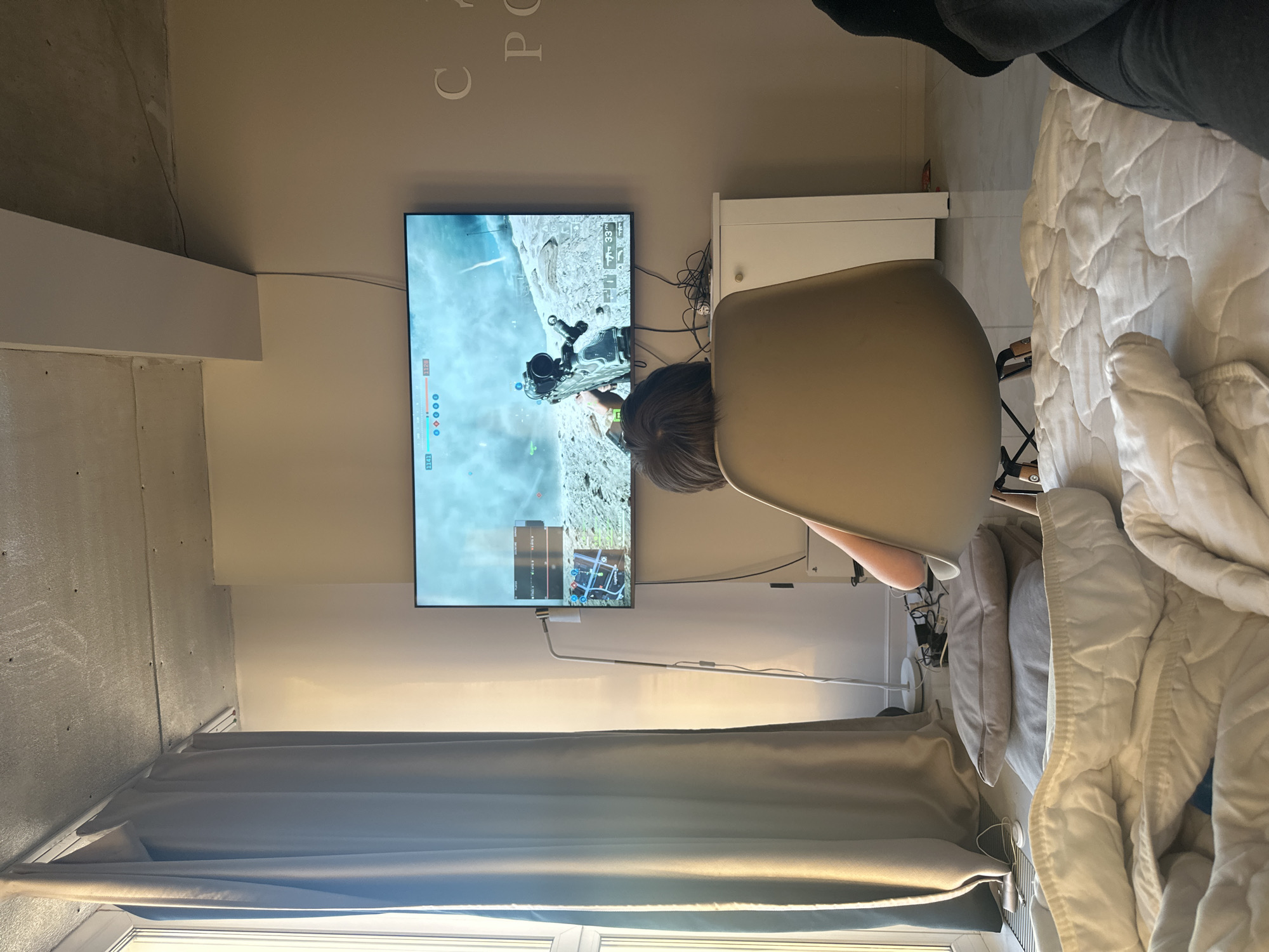 This image depicts an indoor scene, likely a bedroom or living space, in Moscow, Russia. A person, viewed from behind with dark hair, is seated in a light-colored, modern chair, intently playing a video game on a large flat-screen television. The game displayed is a first-person shooter or action game, showing a weapon, a rugged environment, and a user interface with numerical readouts such as "3333," "33," "1150," and "3341," along with blue circular indicators and a mini-map in the top right.
Adjacent to the chair and television, a bed is partially visible on the right, adorned with a textured white quilted comforter and pillows. A white cabinet or bedside table stands to the right of the TV, behind which a cluster of cables and wires is visible. The room's architecture includes an exposed concrete ceiling or wall section on the far left, contrasted with smooth, light-colored walls. Dark curtains hang below a window, allowing ambient light to illuminate the space, suggesting it is daytime. A white floor lamp is partially visible near the bed. On the wall above the television, faint embossed or painted letters, including "C", "P", and "O", can be discerned. The scene conveys a focused gaming session within a contemporary setting.