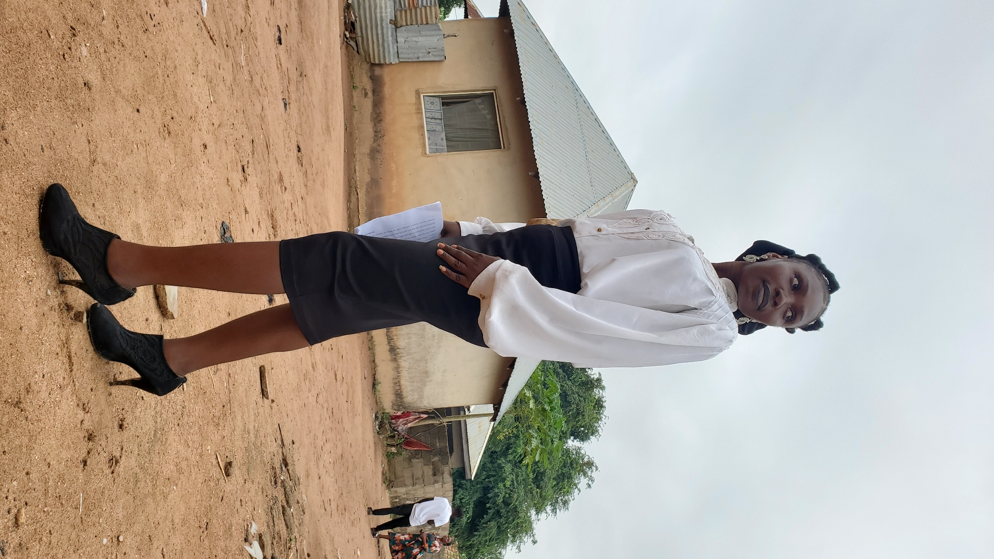 A woman in a white blouse and black skirt stands in a sandy area. She is wearing black heeled boots and holding a piece of paper. In the background, there is a modest house with a corrugated metal roof, and some other buildings and trees. Two other figures are visible in the distance. The sky is overcast. The location is Jalingo, Nigeria.