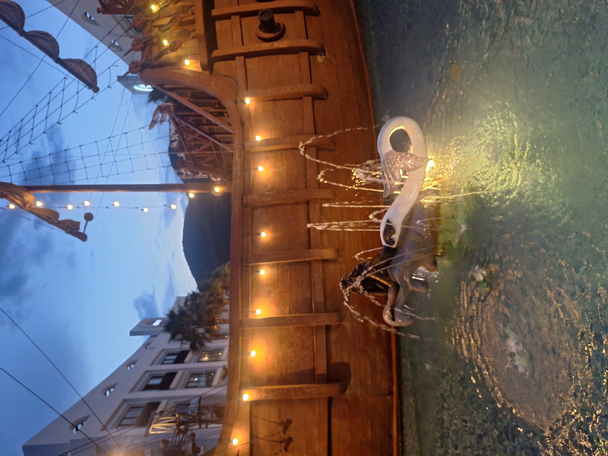 The image depicts a nighttime scene at the VinWonders Nha Trang amusement park on Hon Tre Island, Nha Trang, Vietnam. The foreground features a fountain with water jets spraying from sculpted figures, likely part of a themed water attraction. One sculpted element appears to be a white, abstract shape with metallic accents, while another is a dark, possibly bronze, figure with a mermaid-like tail. The water in the fountain is illuminated by underwater lights, creating a shimmering effect.
Behind the fountain, a large, wooden, pirate ship-themed structure dominates the midground. This structure is adorned with numerous warm-toned string lights, highlighting its textured wooden planks and architectural details, which include railings and what appears to be a ship's mast and rigging in the background.
In the background, a modern, multi-story building with a white facade and dark-paned windows is visible, suggesting the presence of hotels or other resort facilities within the amusement park. Palm trees are scattered between the building and the pirate ship structure, adding to the tropical ambiance. The sky above is a deep twilight blue, with some clouds present, indicating the time of day is after sunset. The overall impression is one of a lively, illuminated, and themed entertainment area.