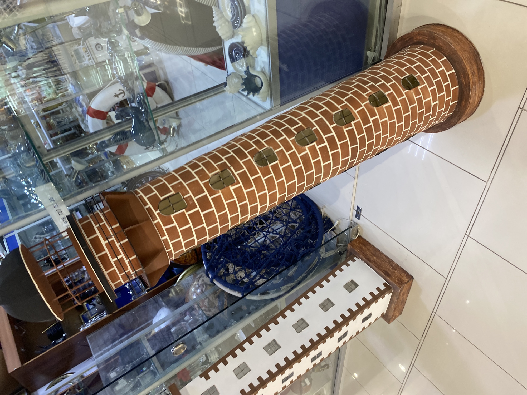 The image displays an indoor scene, likely a shop selling decorative items, featuring two prominent models. Dominating the foreground is a large, cylindrical model resembling a brick lighthouse, positioned diagonally across the frame. It is painted with a detailed brown and white brick pattern and features multiple small, rectangular window cutouts along its body. The top section of the lighthouse model includes a brown observation platform with railings and a dark, possibly domed, cap.
Adjacent to and partially beneath the lighthouse model is a longer, rectangular model, possibly representing a building or fort. This model is white with brown crenellated edges and small square window cutouts. Both models rest near or on glass display cases.
Inside one of the display cases, a white lifebuoy with distinct red stripes and a black anchor symbol is clearly visible. Other nautical-themed decorations and various merchandise, somewhat indistinct, are also present within the glass cases. A blue, net-like object, possibly a decorative fishing net or part of a sporting item, lies on a surface beneath a display case. The floor is tiled with large, light-colored rectangular tiles, suggesting a modern retail environment. The bright, even lighting indicates an indoor setting, likely illuminated by artificial lights. No people are visible, and the scene depicts a static display of goods. Some blurry, partially obscured numbers ("978 727 5..." and "234") are faintly discernible on a tag or reflection in the background glass, but no fully legible text is present.