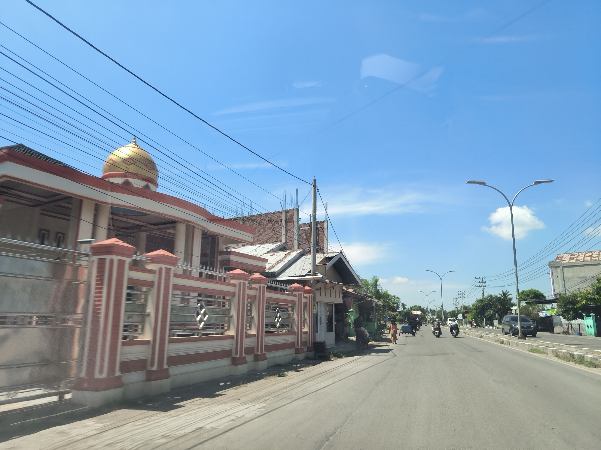 The image depicts a street in Lhokseumawe, Indonesia. The view is from a vehicle traveling on the right side of the road. On the left is a building with a golden dome and an ornate red and white fence. Further down are small shops with people and carts. In the distance, there is a vehicle, a motorcyclist, and some pedestrians. The sky is blue with some clouds. Streetlights line the right side of the road.