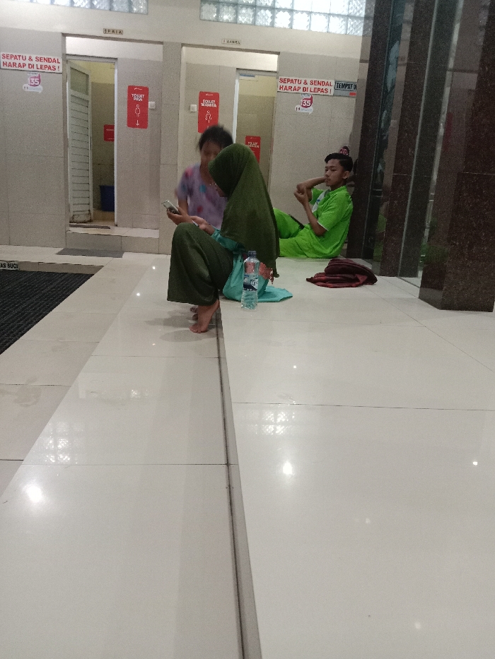 This image captures an indoor public area in Cabean, Indonesia, adjacent to restroom facilities. Three individuals are present.

In the foreground, a woman wearing a dark green hijab and loose green clothing sits barefoot on a raised, tiled platform, actively viewing a white smartphone held with both hands. A plastic water bottle with a blue label and a light blue fabric item rest on the platform beside her.

Behind this woman, a second woman, partially obscured and blurred by motion, stands or moves while also viewing a smartphone.

To the right, a man in a bright green long-sleeved shirt, green pants, and a red-patterned cap is seated on the lower floor, leaning against a dark, vertical structural element. He holds his left ankle with his left hand. A folded maroon fabric item lies on the floor near his right side.

In the background, two open doorways are visible. The left doorway is labeled "PRIA" (Male) and features a red sign with a male figure and "TOILET PRIA." The right doorway is labeled "WANITA" (Female) and has a red sign with a female figure and "TOILET WANITA." Both doorways display signs instructing "SEPATU & SENDAL HARAP DI LEPAS!" (Please remove shoes & sandals!). A blue bucket is partially visible inside the male restroom.

The environment features a glossy, white-tiled floor reflecting overhead lights. A dark, ribbed entrance mat labeled "MAS SUCI" is positioned on the lower floor near the male restroom entrance. Light-colored walls are visible, topped by a row of glass block windows.