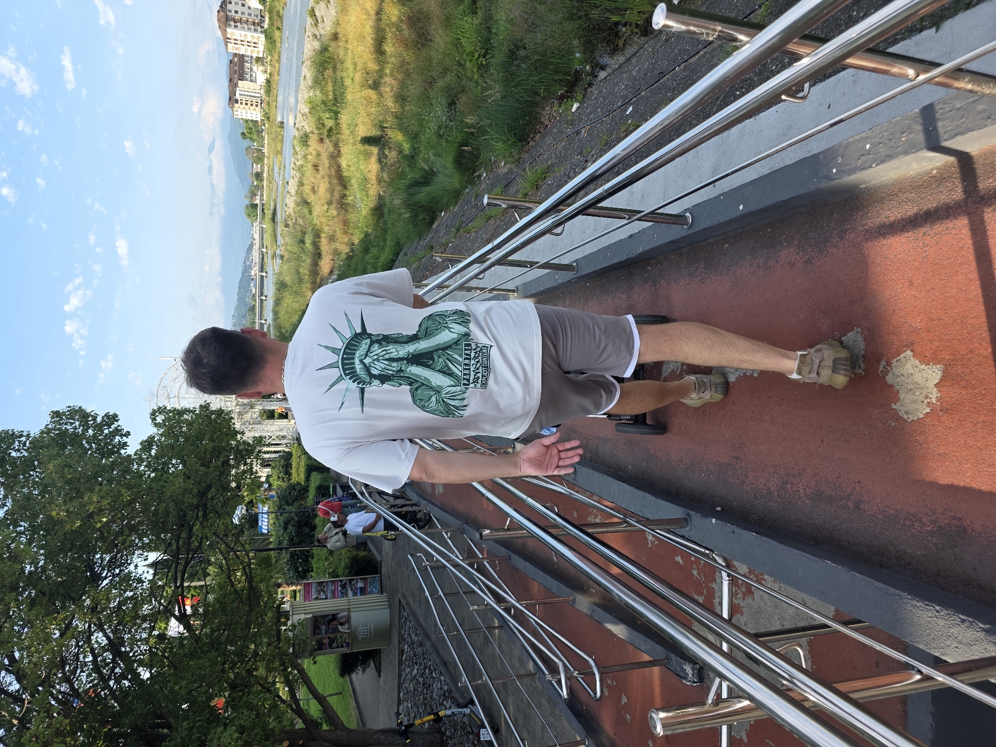 In Sochi, Russia, a person walks away from the camera on a ramp. The person is wearing a white t-shirt with a graphic of the Statue of Liberty covering its eyes, and gray shorts. The person is pushing a small black wheeled cart. The ramp is red and bordered by polished metal railings. To the left of the ramp are trees, a kiosk, and other people. In the background, there is a river, buildings, and mountains under a blue sky with clouds.
