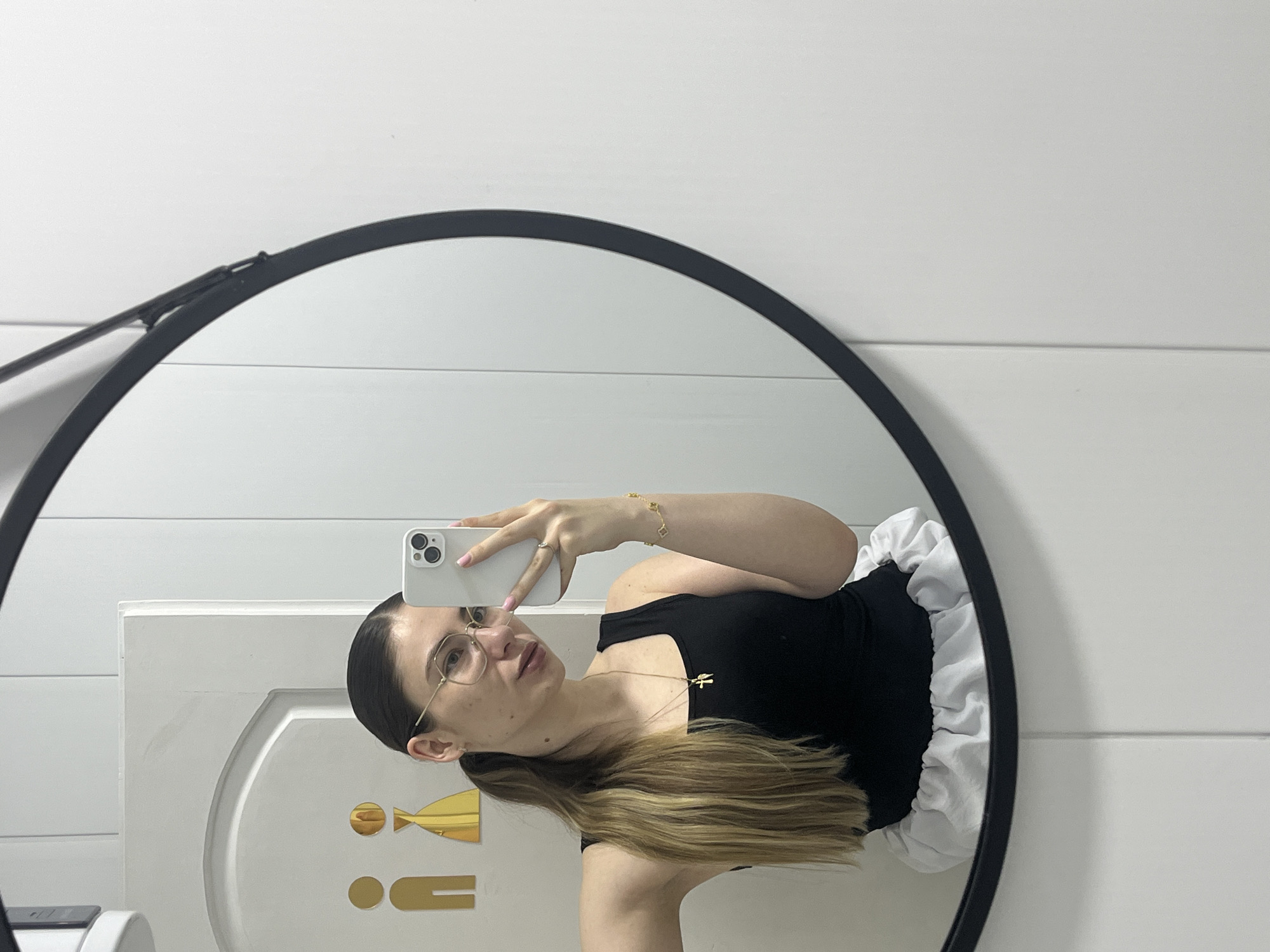 A young woman is taking a mirror selfie in what appears to be a bathroom. She is wearing a black tank top and a white ruffled skirt or underskirt. She has her hair pulled back in a ponytail and is wearing glasses and jewelry, including a necklace with a cross pendant and a bracelet. A phone is visible in her hand as she takes the picture. In the background, reflected in the mirror, is a door with two gender symbols on it, one for men and one for women, indicating a restroom. The walls are white and paneled.