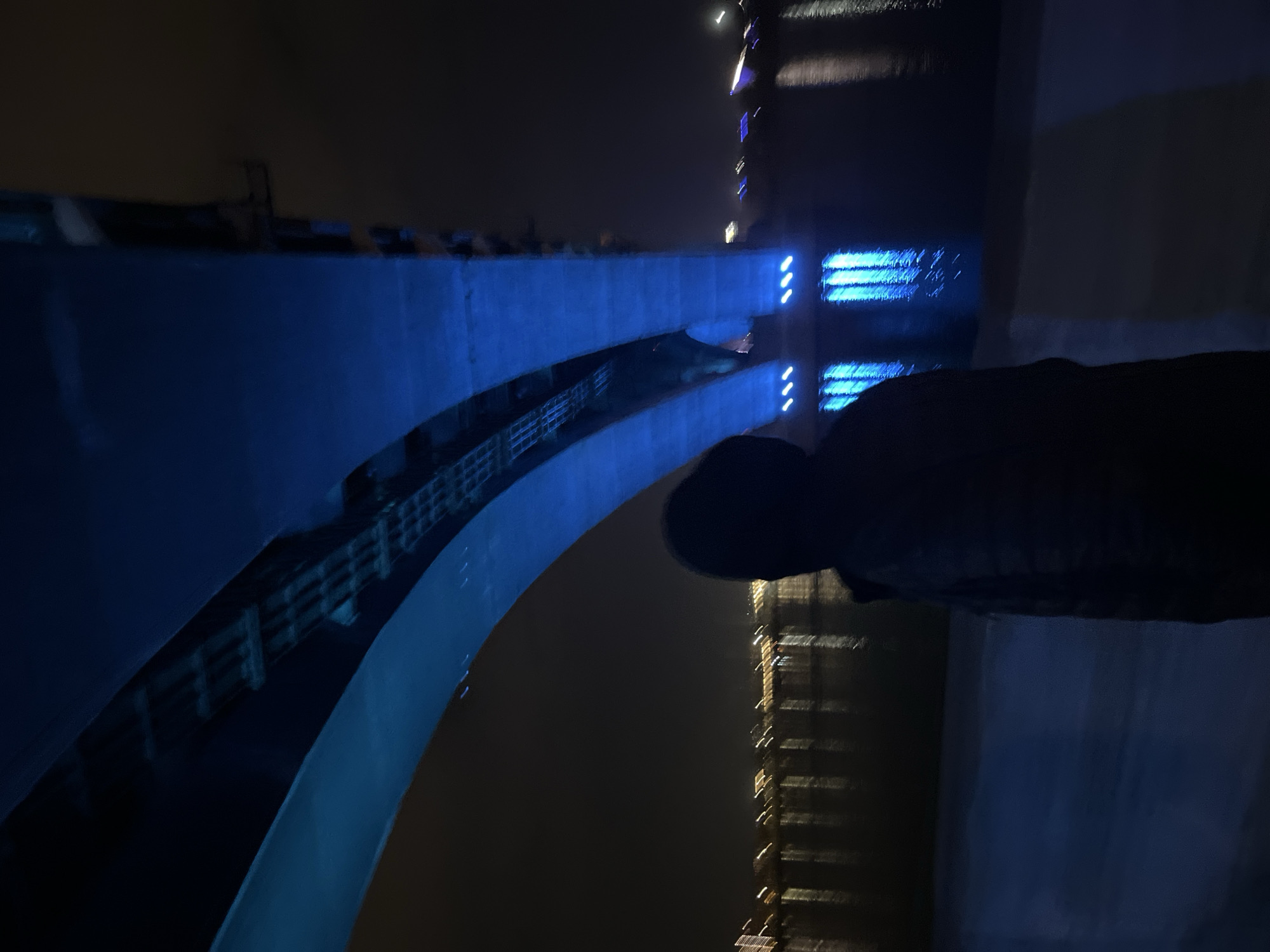 The image depicts a bridge at night in Krasnoyarsk, Russia. A figure stands in the foreground on what seems to be an adjacent structure, partially obscuring the view. The bridge is illuminated with blue lights, contrasting with the dark water reflecting distant lights. A tall building with lights can be seen in the background. The overall scene suggests a nighttime urban setting along the Yenisey River.