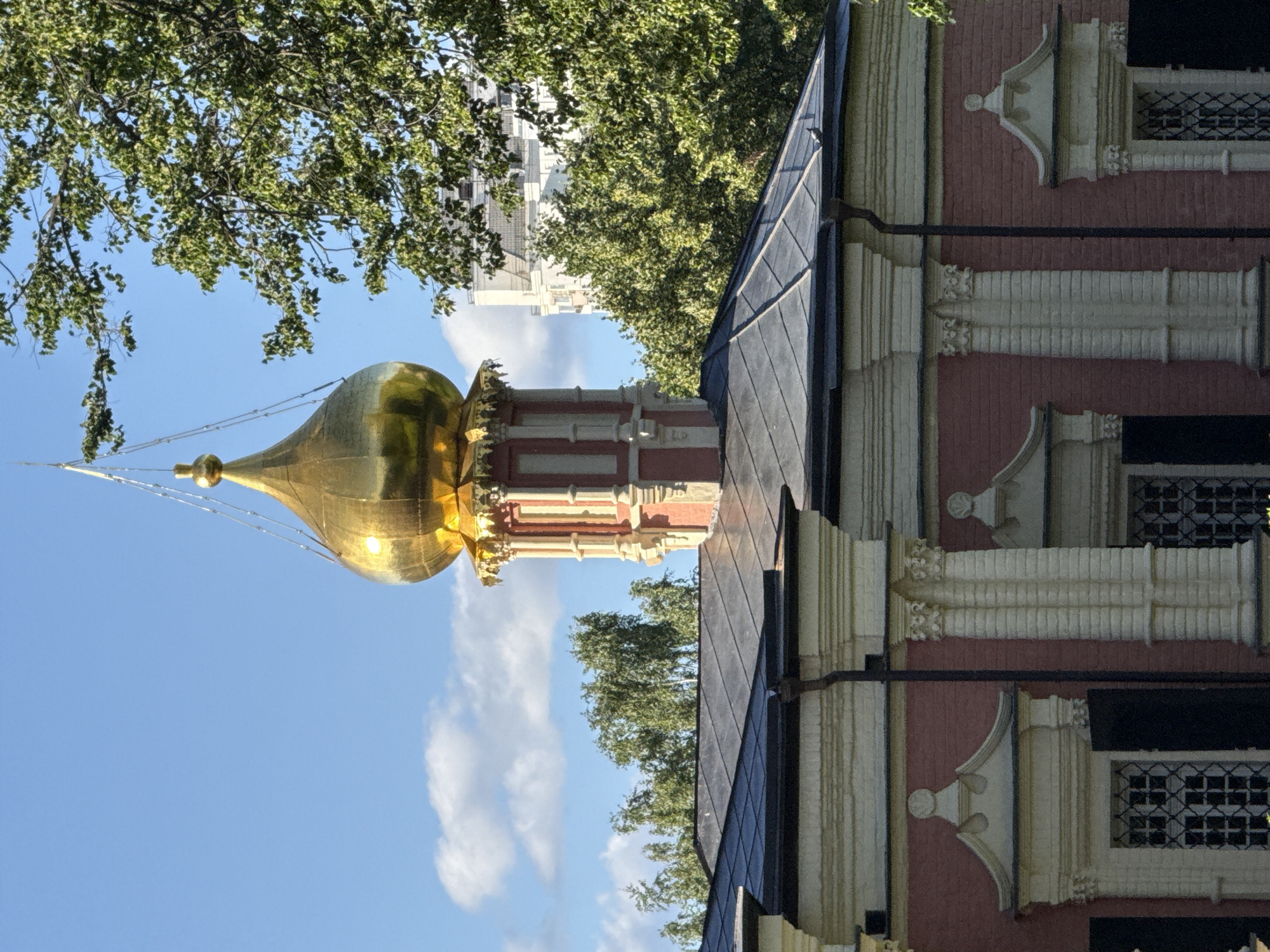 This is a close-up, vertically oriented photograph of a portion of a historical building, likely a church, with a golden dome in the background. The building's facade is adorned with intricate architectural details, featuring alternating bands of red brick and decorative white stone elements. These elements include pilasters, cornices, and decorative window surrounds, suggesting a classical or baroque architectural style. The roof is covered with dark, likely metal, shingles.
In the background, a bright golden dome, characteristic of Russian Orthodox churches, is visible, topped with a cross. The dome is surrounded by the green foliage of trees, suggesting a setting with mature vegetation. A clear blue sky with scattered white clouds serves as the backdrop, indicating fair weather. Hints of modern buildings are visible behind the trees, suggesting an urban environment.
The time of day appears to be daytime, likely late morning or early afternoon, given the bright sunlight illuminating the golden dome and casting subtle shadows. There is no indication of any people or specific activity taking place in the image. The overall mood is one of architectural grandeur and serene natural elements. No text is visible in the image. The architectural style and the golden dome strongly suggest a location within Moscow, Russia.