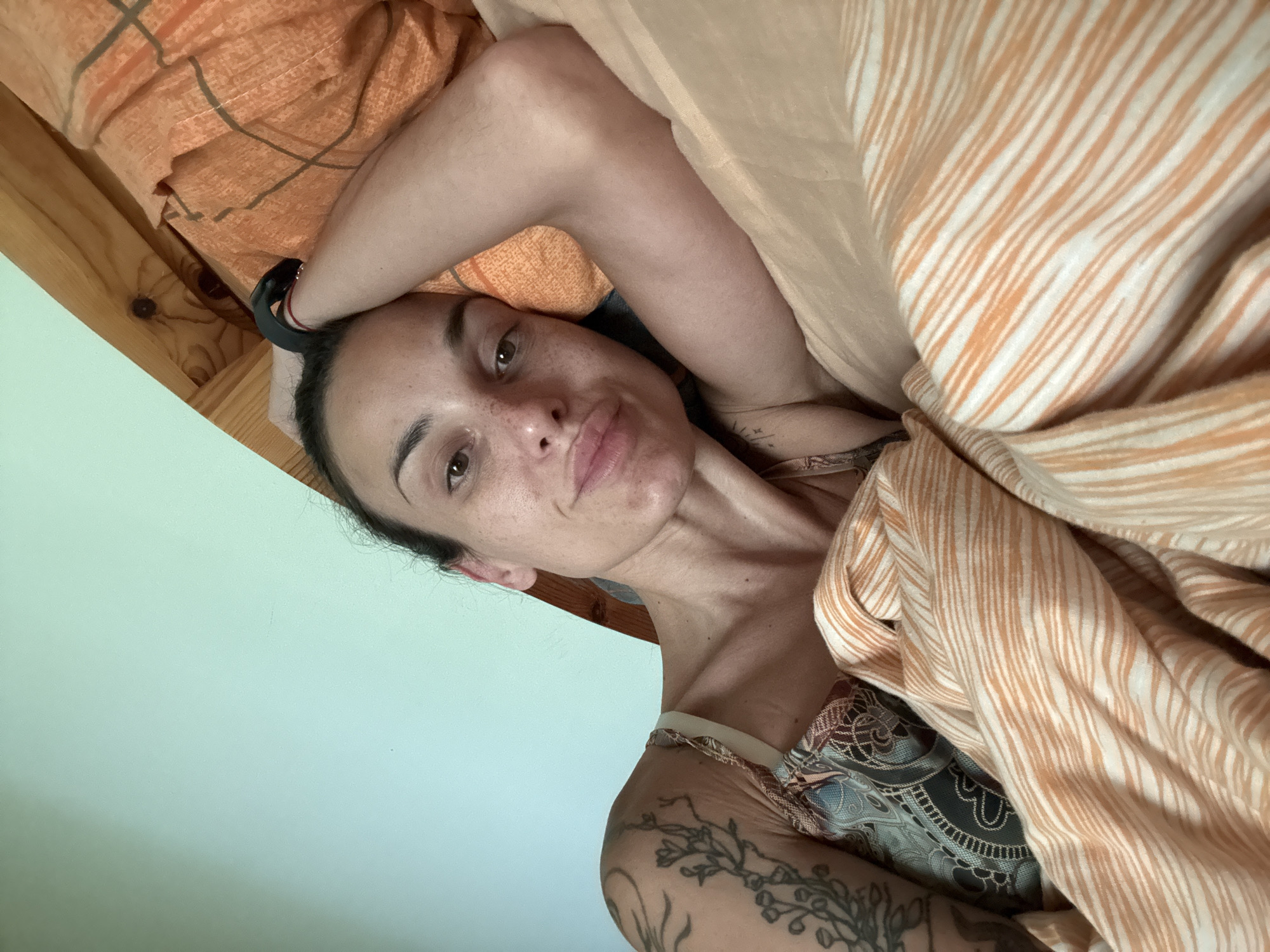 A woman lies in bed, looking at the camera. Her left arm is bent and resting above her head, with her hand behind her head. Her right arm is partially visible, bent at the elbow and resting beside her body. She has dark hair pulled back from her face and appears to have freckles. She is wearing a patterned top. The bed is covered with orange and white striped sheets and a light orange blanket. The background shows a light blue wall and wooden headboard. The scene is indoors, likely a bedroom. The image does not contain information to determine the location in Pleven, Bulgaria.