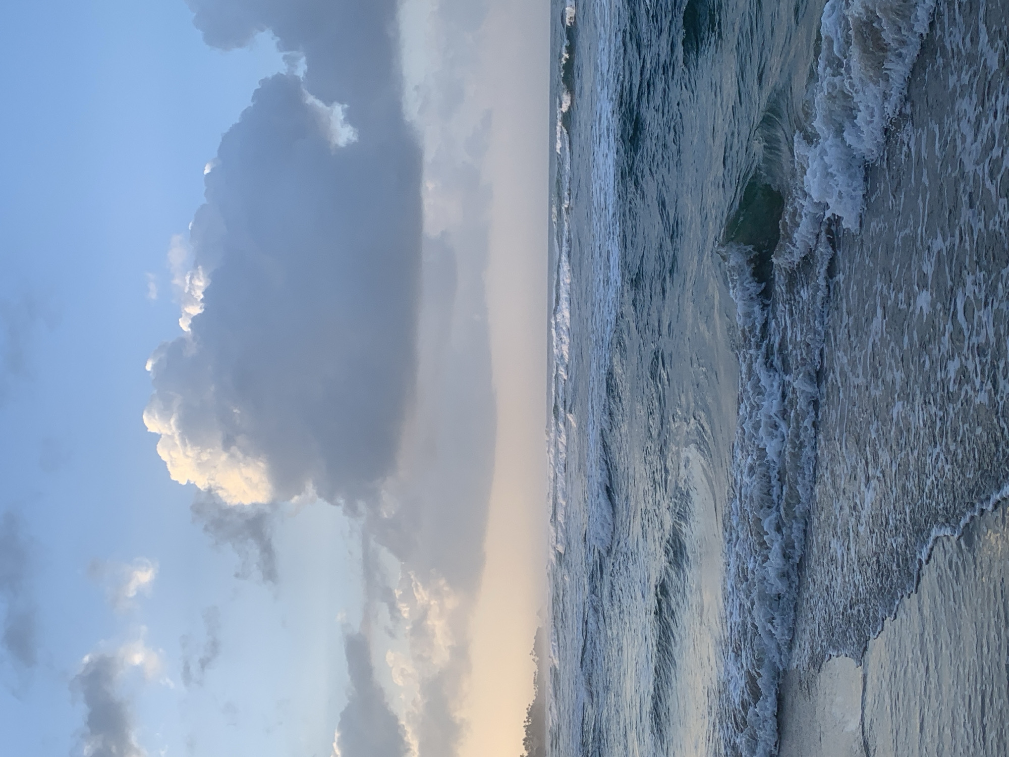 A vertical, eye-level, medium shot captures a turbulent ocean with a sandy shore in Kathaluwa, Sri Lanka. The sky is filled with dramatic, large, white and gray clouds that are illuminated by the soft light of dawn. The ocean is a deep blue-green, with white waves crashing onto the shore and creating foamy patterns in the shallow water. A distant, hazy shoreline with palm trees is visible on the left horizon. The image conveys a sense of movement and the raw power of nature.