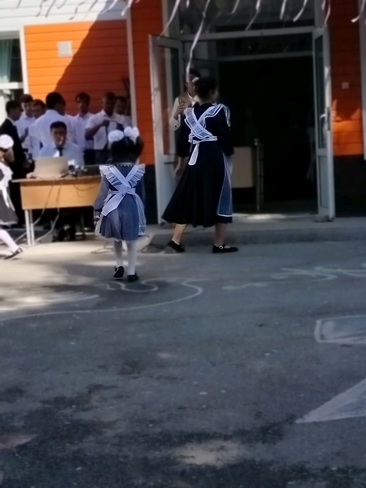 Two young girls in school uniforms, one smaller than the other, are walking on a paved outdoor area. The smaller girl, in a blue dress with a white apron and large white bows in her hair, is facing away from the camera. The slightly larger girl, also in a school uniform with a dark dress and white apron, is walking towards an open doorway. A group of young people in white shirts and dark pants are gathered in the background near a building with orange siding. A wooden desk with a laptop is visible to the left. The ground is a dark asphalt surface with some chalk markings. The scene appears to be at a school in Kölkent, Kazakhstan, possibly during an event or on the first day of school.