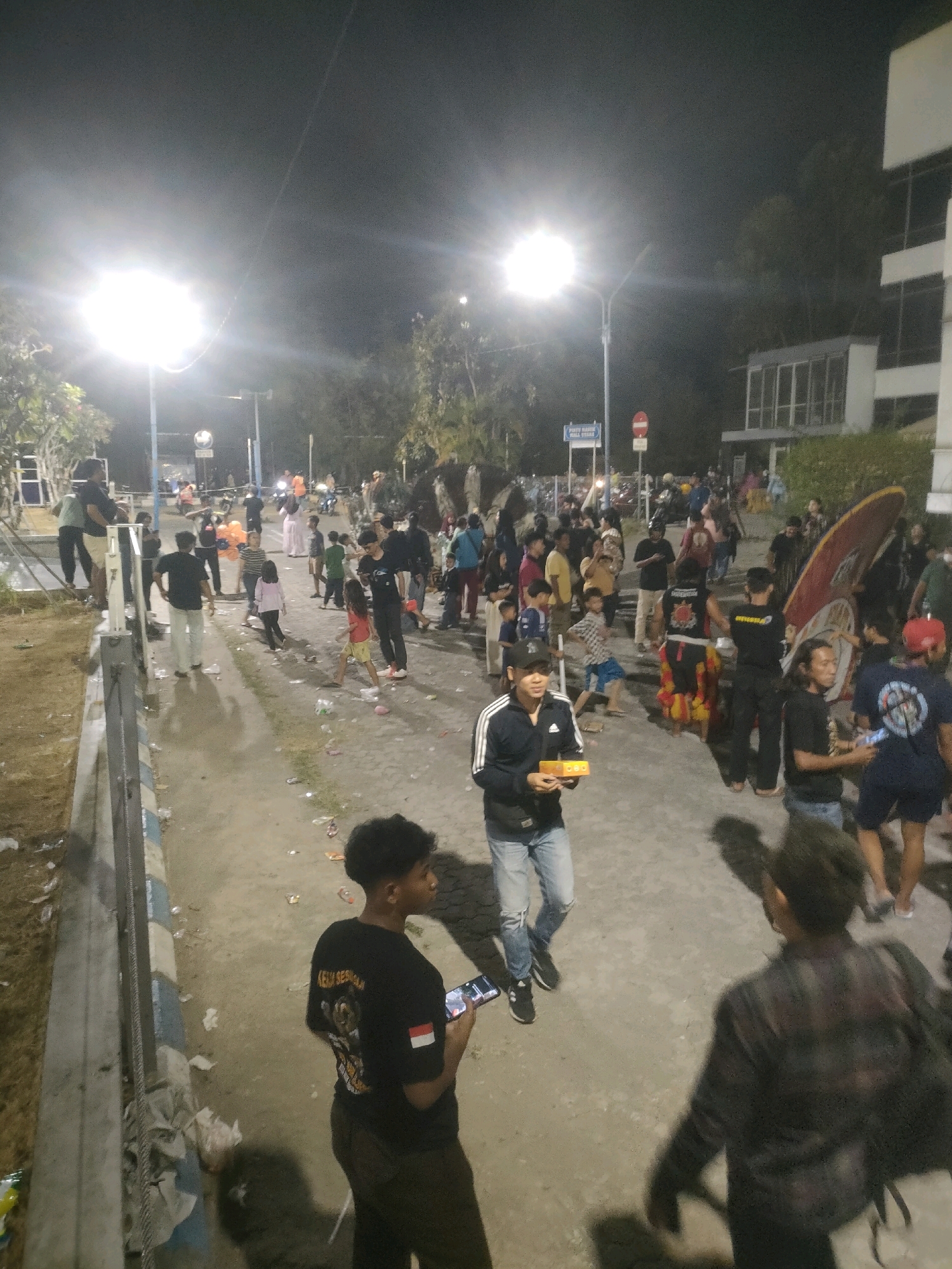 The image depicts an outdoor night scene, likely a public gathering or event, with strong illumination from streetlights. Several individuals are present, engaged in various activities such as walking, conversing, and holding objects. The ground is paved and appears somewhat littered. In the background, buildings and trees are visible, suggesting an urban or semi-urban environment. One man in the foreground is holding a mobile phone and looking to the side, while another in the center foreground is holding a small yellow box. A person in the far right foreground is captured from behind, wearing a plaid shirt. The presence of diverse attire and the general atmosphere suggest a casual social event. The reference to Surabaya, Indonesia, implies this scene is situated within that city.