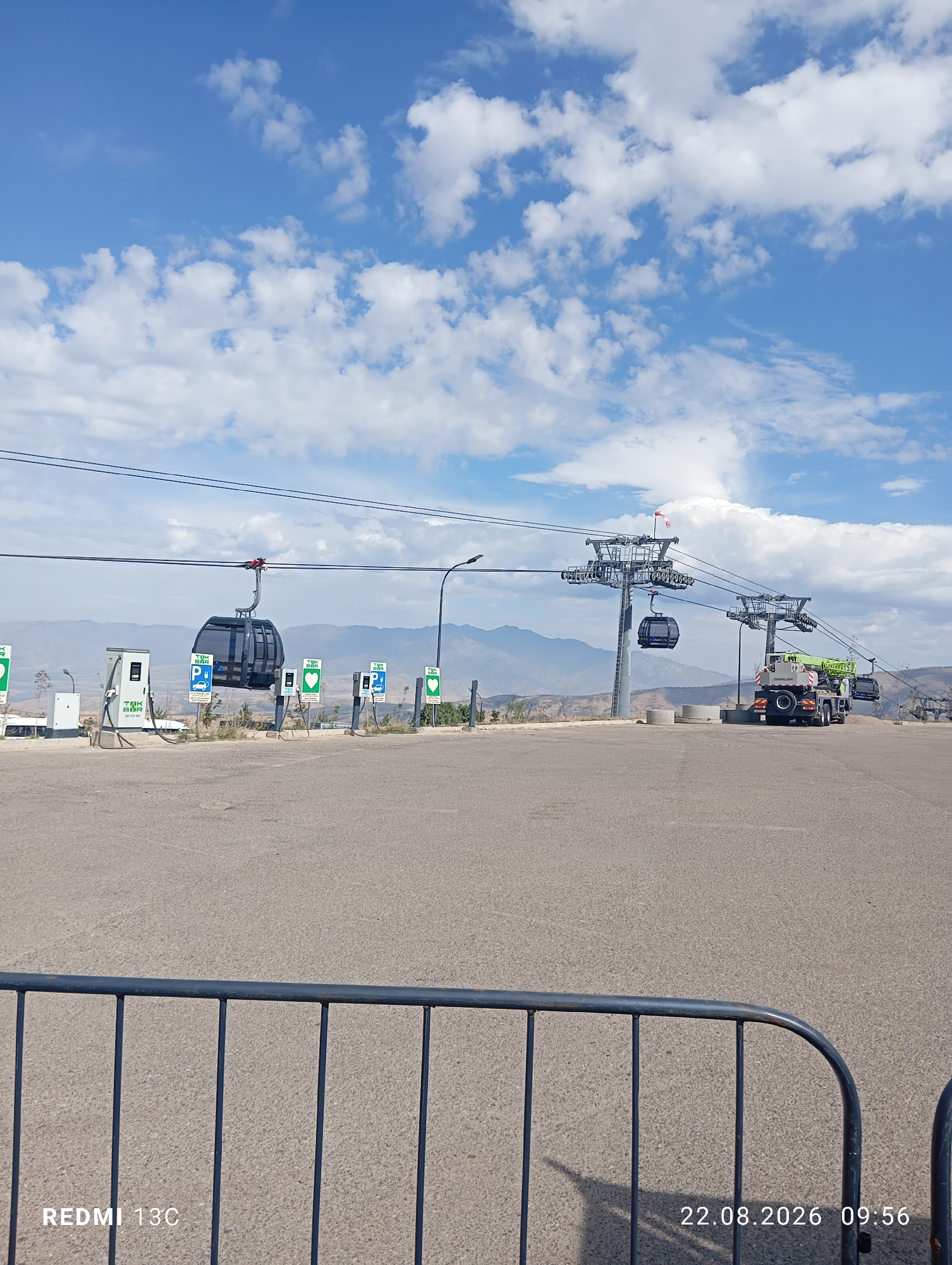 The image depicts a clear sky with scattered clouds over a mountainous landscape, characteristic of the Amirsay ski resort in Uzbekistan. In the foreground, a paved area is visible with several charging stations for electric vehicles, indicated by signs with a lightning bolt symbol and the text "TAK BAR". A cable car, also referred to as a gondola, hangs suspended from wires, suggesting it is part of a ski lift system. Further in the background, a crane is positioned near a cable car support tower, implying construction or maintenance work is in progress. The scene is devoid of people. The date and time stamp "22.08.2026 09:56" and the text "REDMI 13C" are present in the bottom corners of the image.