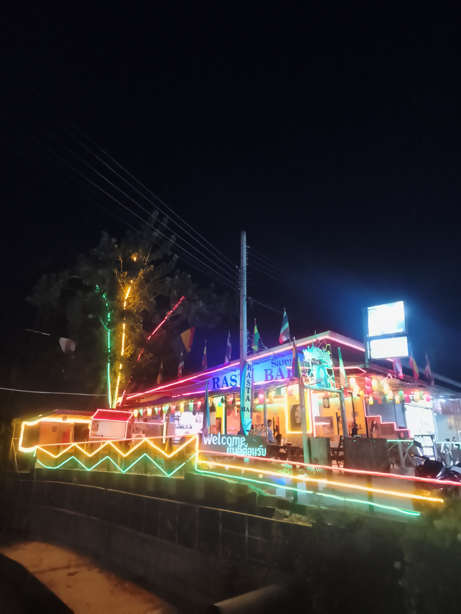 At night, the Samui Rasta Bar is brightly lit with colorful neon lights and festive decorations. The building, located in Baan Chaweng, Thailand, has a sign with "Samui Rasta Bar" written on it. Small flags are displayed along the roof, and a welcome sign is visible near the entrance. Tables and chairs are arranged outside, with several people visible inside the bar. Lights illuminate a nearby tree, and utility poles and wires are visible above the building. Streaks of moving lights suggest passing vehicles.