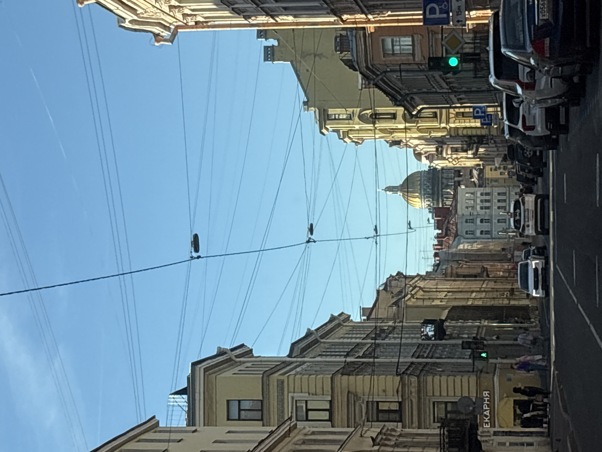 The image depicts a street scene in Saint Petersburg, Russia, likely along Nevsky Prospekt, a prominent avenue known for its historic architecture and St. Isaac's Cathedral, the golden dome of which is visible in the distance. The photograph captures a sunny day with a clear blue sky.
On either side of the street are tall, multi-story buildings with classical architectural features, painted in muted yellow and ochre tones. The buildings have numerous windows, some with dark shutters and balconies. Overhead, a complex network of electrical wires and traffic lights crisscrosses the sky.
In the foreground, cars are driving on the asphalt road, with traffic lights indicating a green signal. Several people are walking on the sidewalk on the left side of the street. One building has a sign with Cyrillic lettering that reads "ЕКАРНЯ," suggesting a shop or business.
The overall impression is one of a bustling urban environment with a blend of historical grandeur and modern activity, characteristic of Saint Petersburg's city center.