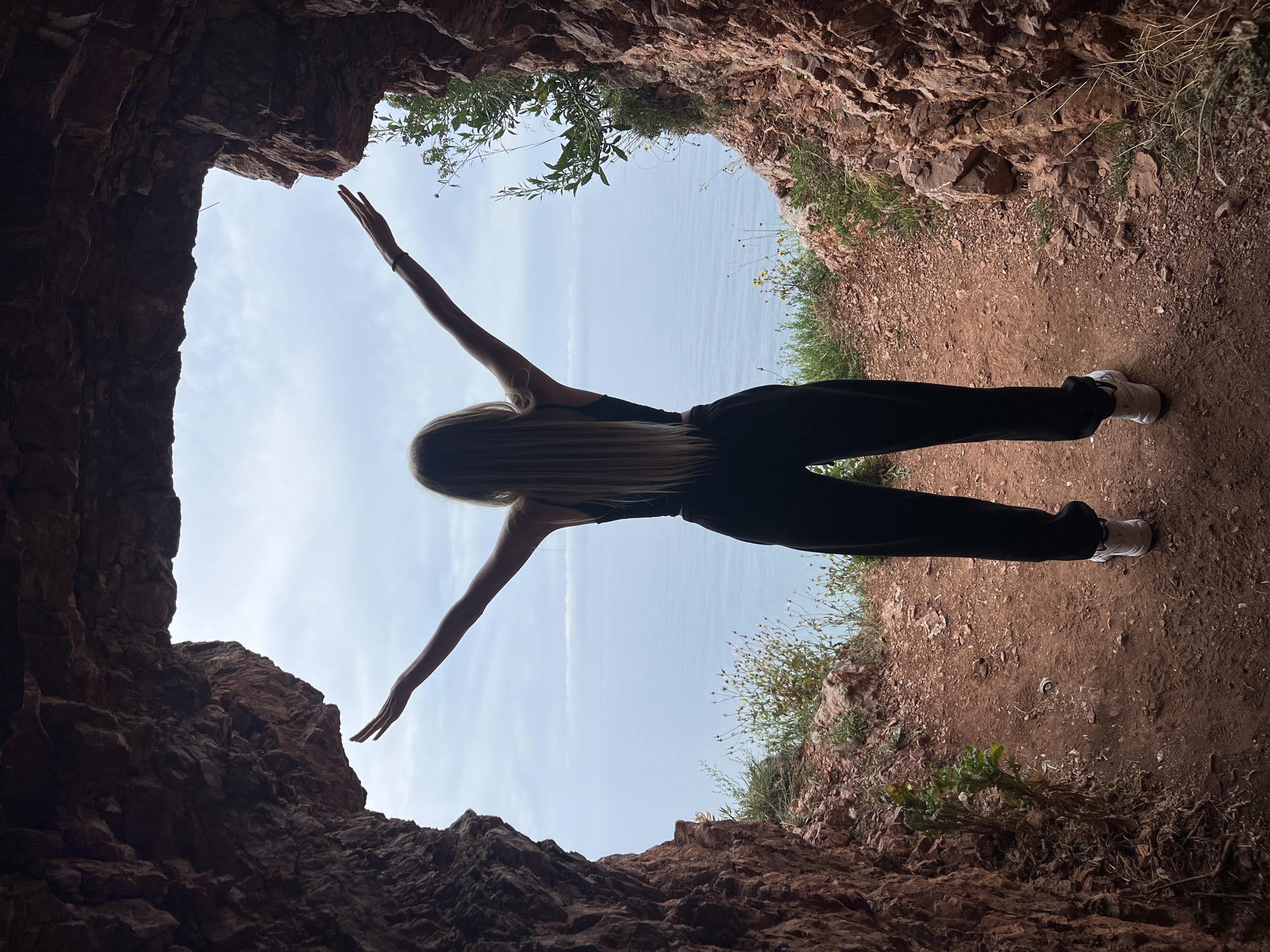 A vertical, low-angle photograph depicts a woman standing at the entrance of a cave. The woman is wearing black pants and a black top. Her arms are extended outwards in a cruciform pose. The cave entrance frames a view of a body of water, likely the Adriatic Sea. The interior of the cave is rocky and earthen, with sparse vegetation visible around the opening. The location is likely near the coast of Petrovac centar, Montenegro, given the sea visible through the opening.