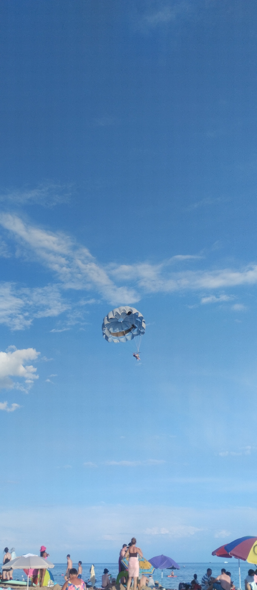 A parasailer is suspended in a bright blue sky with scattered white clouds above a beach scene in Cholpon-Ata, Kyrgyzstan. The parasail is silver and white, with two people visible in the harness below. The beach is populated with numerous individuals, some standing and others sitting under colorful beach umbrellas, suggesting a recreational area. The water of Lake Issyk-Kul is visible in the background. The overall scene captures a sunny day at a popular tourist destination.