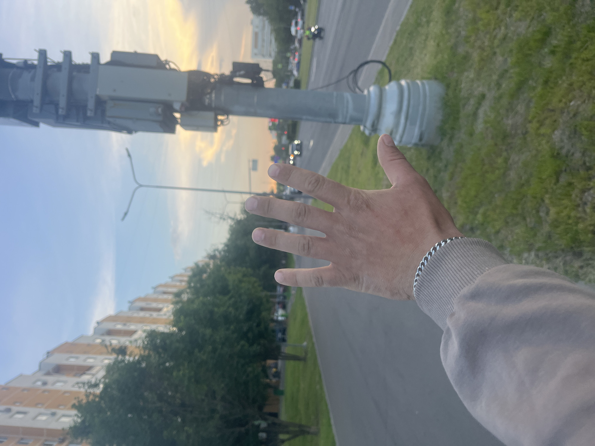 In Moscow, Russia, a person's hand with a chain bracelet is held up, blocking the view of a street with cars and buildings. The street is lined with trees and a sidewalk. A lamppost with electrical equipment is prominent in the foreground, and apartment buildings are visible in the background. The sky is overcast with hints of yellow and orange from what appears to be a sunset or sunrise.