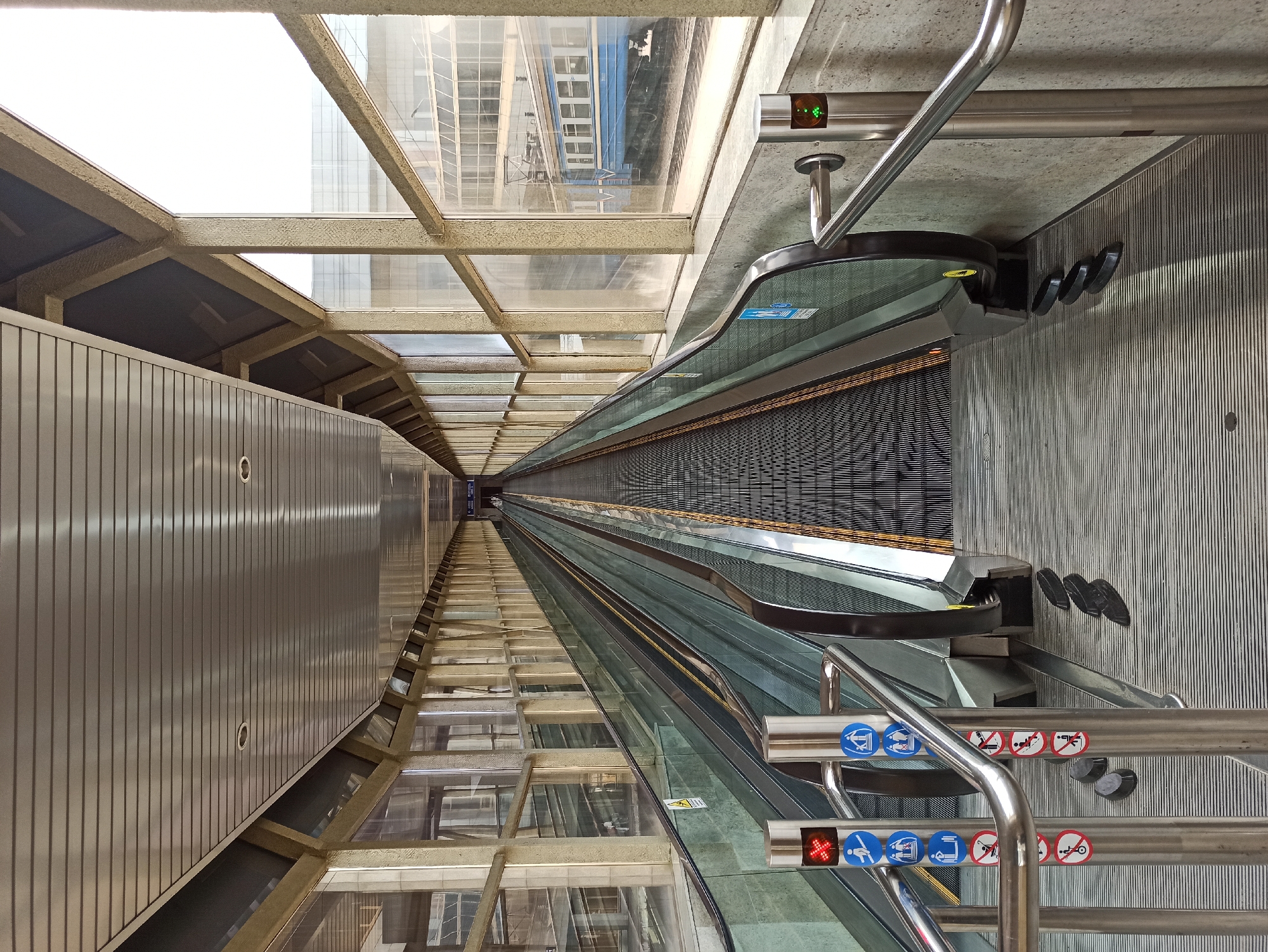The image shows a long, indoor moving walkway at an elevated level, with escalators on either side. The walkway is enclosed by large windows that look out onto a city scene with buildings and a blue and white train on tracks. Several signs with international symbols are attached to metal posts along the sides of the walkway, indicating restrictions and rules. There are no people visible on the walkway itself. The architecture appears to be utilitarian and modern, with metal ceilings and concrete walls. The overall impression is one of an efficient, if somewhat stark, transportation hub. This scene is located at the Minsk National Airport (MSQ) in Minsk, Belarus, specifically in the pedestrian bridge connecting Terminal 1 to the car park.