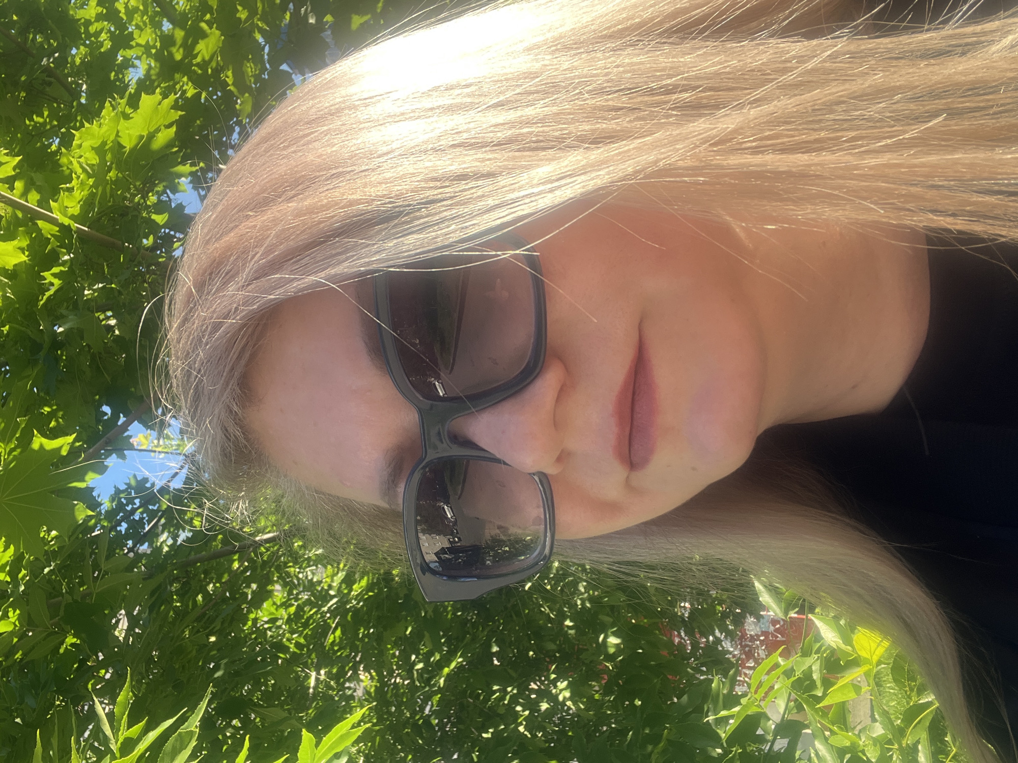 A close-up, right-facing profile shot captures a woman with blonde hair and sunglasses. She is outdoors, with lush green foliage and a bright blue sky filling the background. The sunglasses reflect a street scene with buildings and cars, indicating an urban or suburban setting. The lighting suggests it is daytime, with sunlight illuminating her hair and face. There are no specific identifying landmarks visible that would pinpoint a location within Izhevsk, Russia.