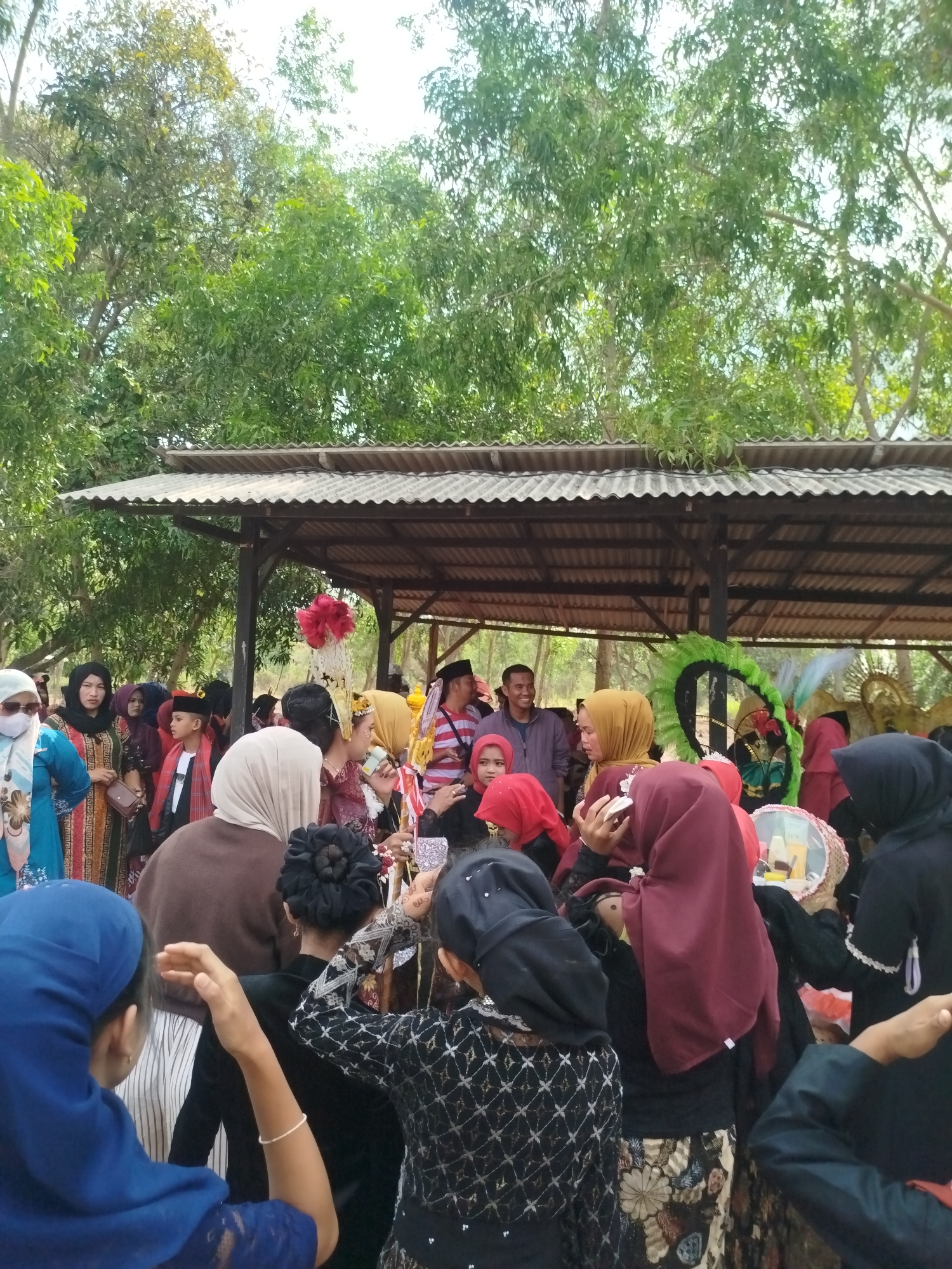 The image depicts an outdoor gathering of people in Jambu, Indonesia, under a covered structure. A crowd, predominantly women dressed in various colored hijabs and traditional clothing, is gathered around a central area. Several individuals are wearing ornate headdresses, suggesting a celebration or cultural event. In the background, lush green trees form a natural canopy, and the sky is visible through the leaves, indicating it is daytime. The overall scene conveys a sense of community and festivity.