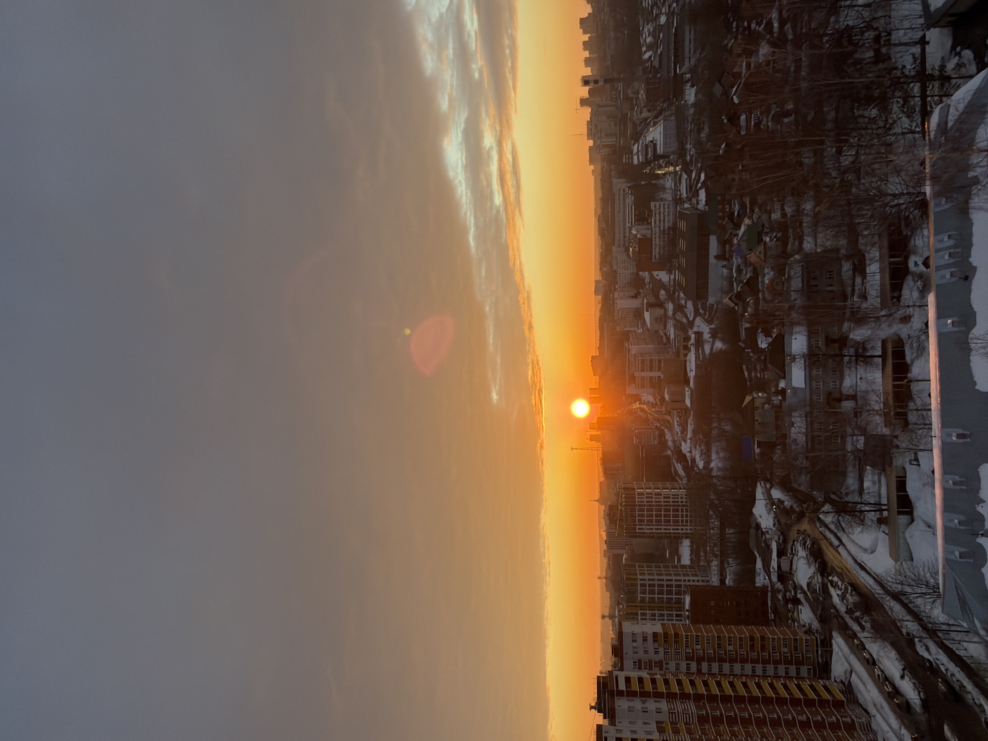 An elevated, wide-angle photograph captures a panoramic cityscape of Izhevsk, Russia, during what appears to be either sunrise or sunset in winter. The sky dominates the upper two-thirds of the image, transitioning from a dull gray-blue at the zenith to an intense orange-yellow at the horizon. The sun, a bright, distinct orange-yellow disk, is positioned low on the horizon, slightly right of the center. A prominent reddish-orange lens flare artifact is present in the upper-left quadrant of the sky. Sparse, thin clouds are visible, some illuminated by the sun's warm light, others appearing as dark silhouettes.
The cityscape extends from the midground to the distant horizon. On the far left, a multi-story residential building with reddish-brown, white, and yellow facades is prominently visible. Further into the midground, numerous other high-rise buildings, some with modern, gridded architectural styles, are intermingled with smaller, older structures. The ground throughout the visible urban area is extensively covered in snow. Roads, dark against the white snow, snake through the city, with several parked vehicles visible along them and in a parking lot on the left side.
A distinctive golden-domed spire, consistent with St. Michael's Cathedral, is identifiable on the distant horizon, slightly left of the sun, confirming the location as Izhevsk. Other industrial and residential structures, including several prominent towers and smokestacks, form the urban tapestry, largely silhouetted against the vibrant sky. Deciduous trees, bare of leaves, are scattered throughout, particularly in clusters between buildings and along snow-covered slopes. The immediate foreground features the snow-covered rooftop of a building, reflecting the ambient light. No individuals or specific actions are discernible within the frame. The scene is still and bathed in the low, warm light of the golden hour, contrasting with the cold, snow-covered environment.