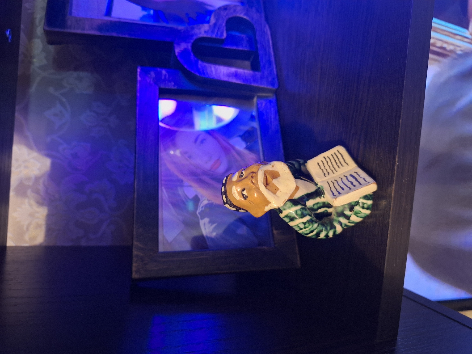 The image displays an indoor scene characterized by dark wooden elements and striking blue artificial lighting. In the foreground, a ceramic figurine of a man is affixed to a dark wooden column or frame, positioned as if reading an open book. The figurine depicts an older man with light brown skin, a white beard and mustache, and a dark cap. He wears a green and white striped garment, and the book he holds shows stylized wavy lines representing text.
Behind the figurine, part of a wooden structure is visible, which includes a decorative heart-shaped cutout at the top. Below this, there is a rectangular opening, likely a mirror or picture frame, reflecting the face of a young woman. She has long, light-colored hair and appears to be wearing makeup, with her gaze directed somewhat towards the viewer. Another partial reflection, possibly of a torso, is visible in a second, less distinct frame below the first.
The background behind the wooden structure features a patterned wall, possibly wallpaper, with a light floral or damask design that is softly illuminated. The strong blue light source casts a cool, ambient glow over the entire scene, creating distinct shadows and highlights on the wooden surfaces and reflections. The dark wood has a visible grain. The setting appears to be a decorative display within a room, potentially in Moscow, Russia, as per the provided context. There is no legible text present; the book in the figurine's hands features decorative lines mimicking writing.