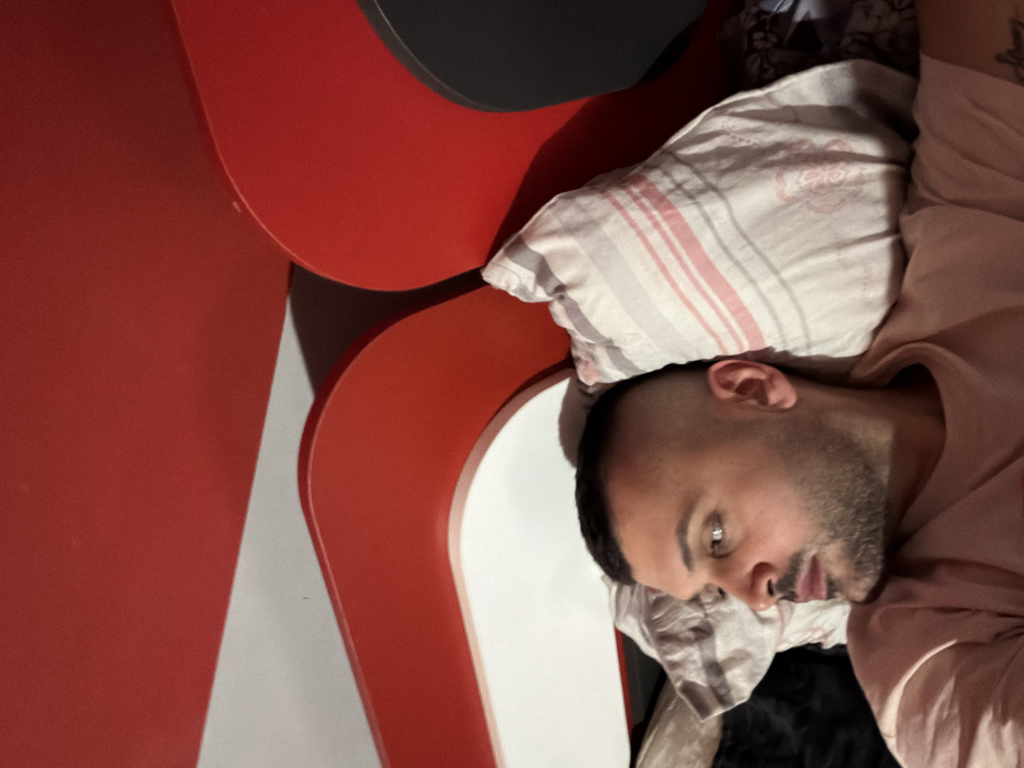 A male individual is observed lying on his back in an indoor setting located in Opanets, Bulgaria. The subject, an adult male with short dark hair in a high fade style and a trimmed beard, is wearing a light-colored, possibly pink, short-sleeved t-shirt. He is resting his head on a pillow with faint grey and pinkish-red stripes. His head is turned to the right, and his gaze is directed slightly upwards and to his left. The background consists of a wall or headboard featuring prominent, overlapping curved decorative elements in solid red and white, with a darker, possibly black, element visible on the far right side of the frame. The lighting suggests an interior environment, casting shadows among the decorative elements.