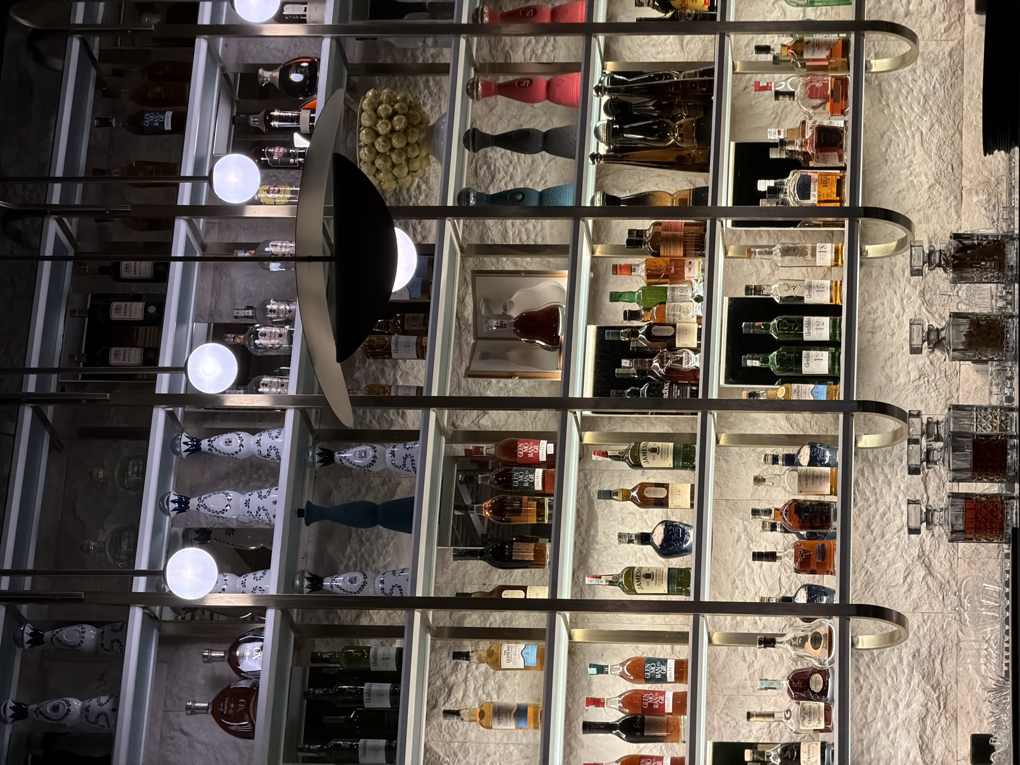 The image shows a liquor cabinet displaying various bottles of alcohol, including Glenfiddich, Glenlivet, Jameson, and Martell, arranged on shelves. The bottles are predominantly arranged on shelves, with some decorative items mixed in. A lighting fixture with round bulbs and a curved design is placed in front of the cabinet. The background appears to be a wall with a textured surface. The image was taken indoors, likely in a bar or restaurant. The city location is not discernible from the image.
