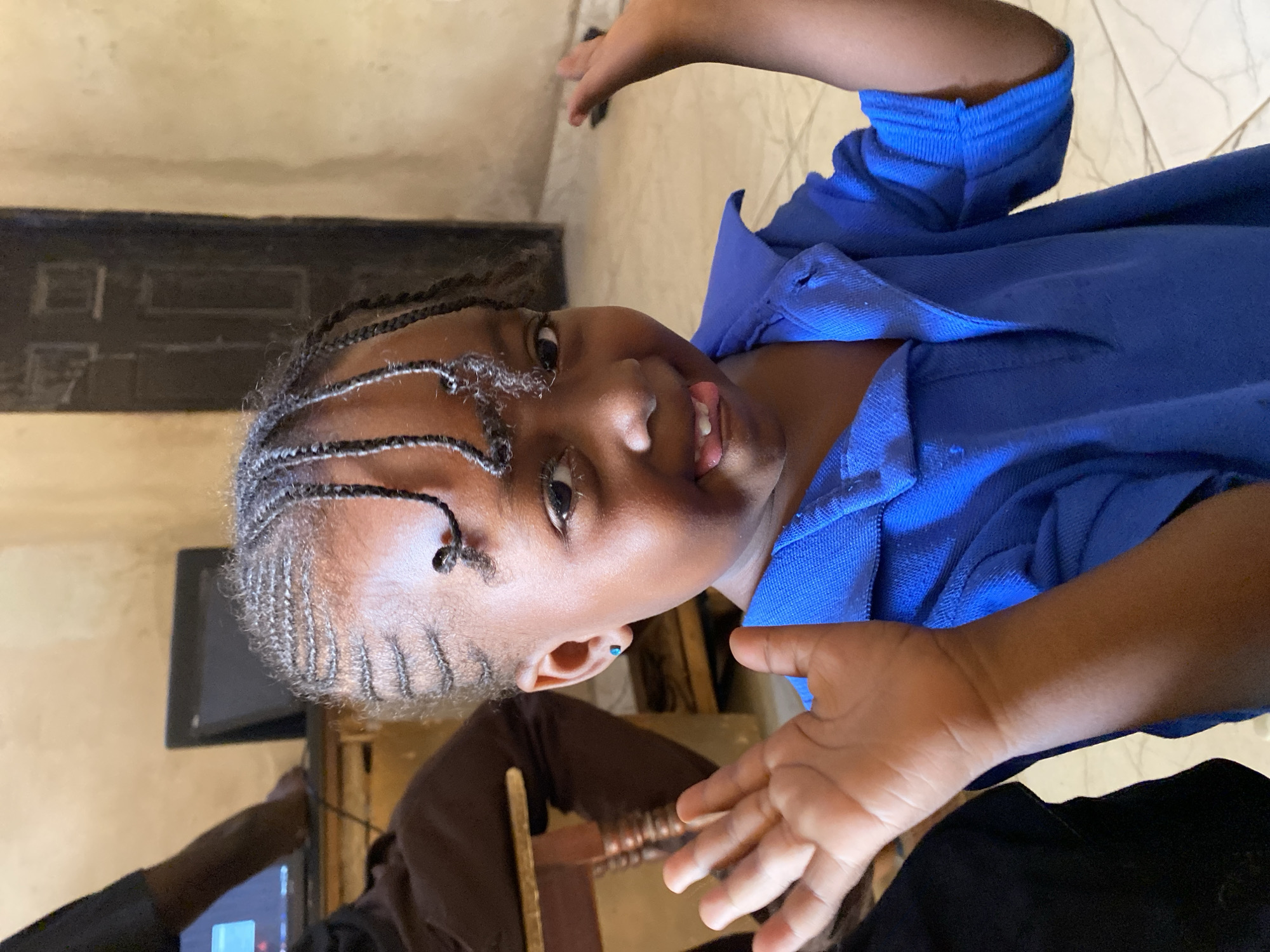 A young Black girl, appearing to be of elementary school age, is the central subject of the image, captured in what seems to be an indoor setting in Kuru Dungus, Nigeria. She is looking directly at the camera with a bright-eyed expression and a playful slight smile, her tongue peeking out. Her hair is styled in numerous small, dark braids that extend from her scalp. She is wearing a vibrant royal blue polo shirt with a collar, and a small, light blue stud earring is visible in her left ear. Her hands are open and extended slightly towards the camera, as if mid-gesture or in playful interaction.
The background indicates an interior space, possibly a home or community center. The walls are a light, warm beige or tan color. The floor is covered with light-colored tiles, possibly white or cream, arranged in a pattern with darker grout lines. In the left background, a dark rectangular object, potentially a door, cabinet, or part of a window, is visible. Below this, the arm of another person, clad in dark clothing, is partially seen, along with a portion of a screen, possibly a television or computer monitor, suggesting another individual is present and engaged with an electronic device. The lighting is diffused, consistent with an indoor environment. There is no visible text in the image.