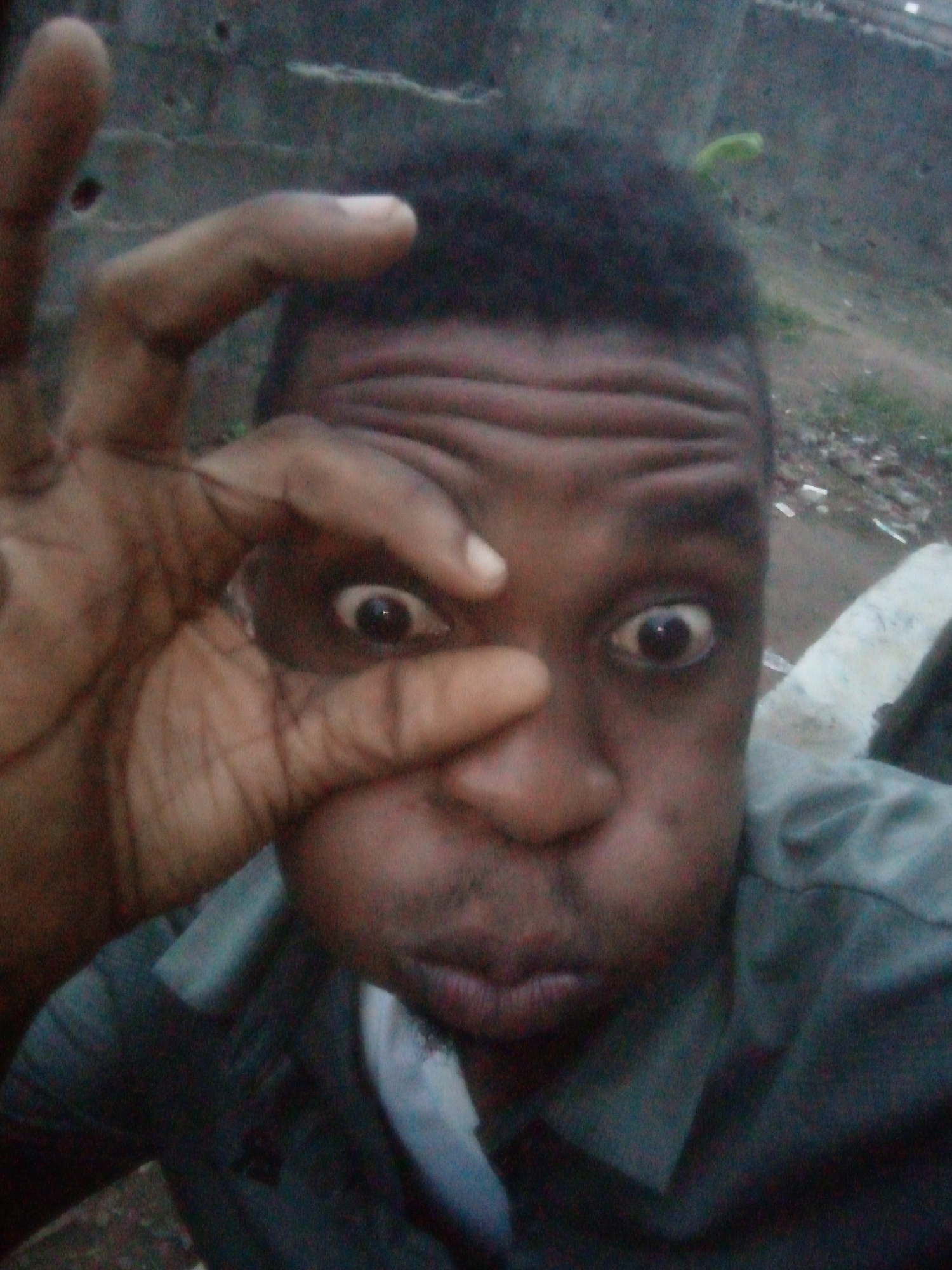 A medium shot captures a person with dark skin and short, dark hair, making a distorted face. Their cheeks are puffed out with air, and their eyes are wide and exaggerated. The person has placed their hand up to their face, with their fingers forming a small circle around one eye, further emphasizing the facial distortion.
The background is blurred and indistinct, appearing to show an exterior environment, possibly a city, with a mix of foliage and scattered debris on the ground. A partial wall or fence is visible in the background as well.